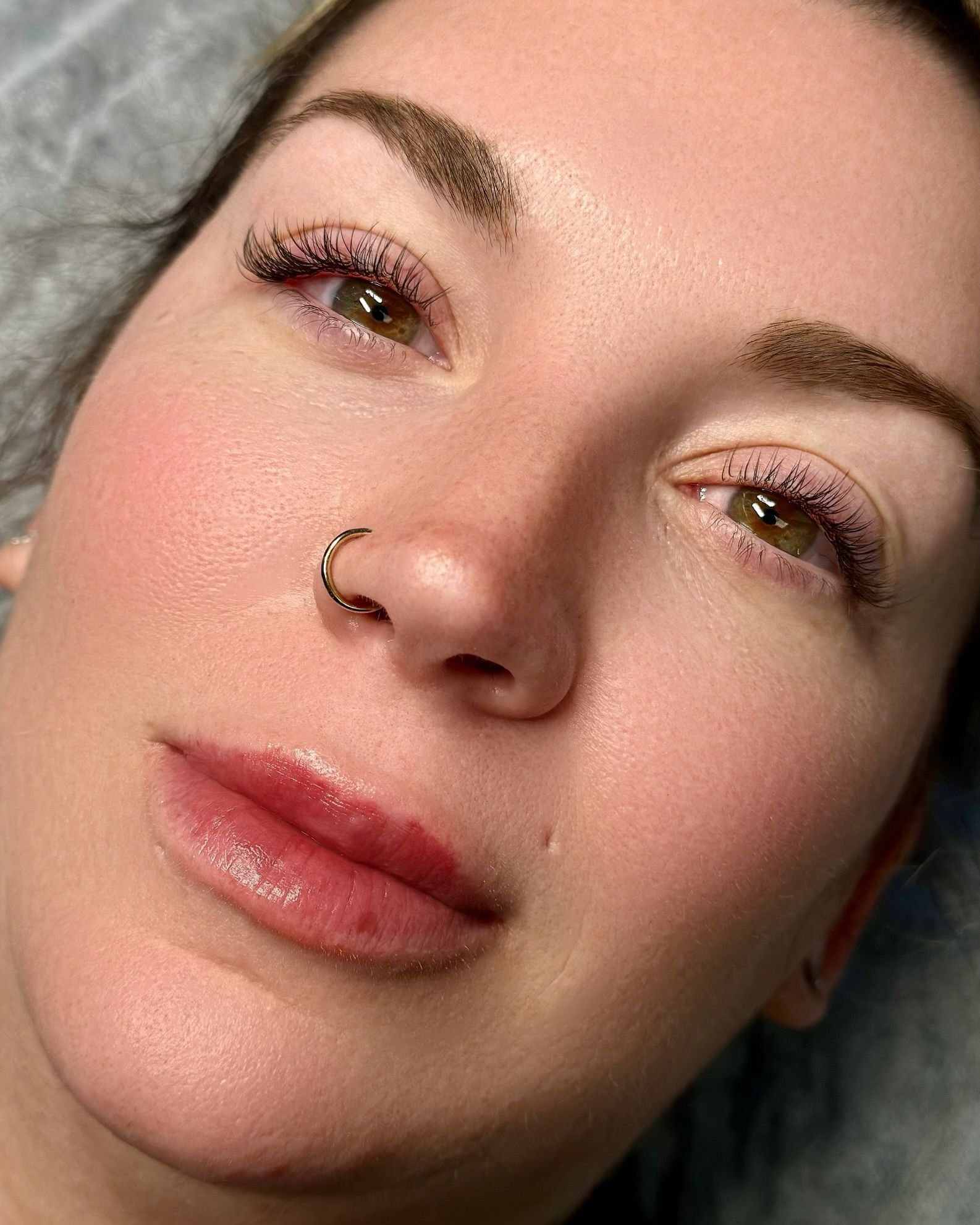 Woman with enhanced eyebrows, eyeliner, and lips. Pink blush on cheeks, nose ring.