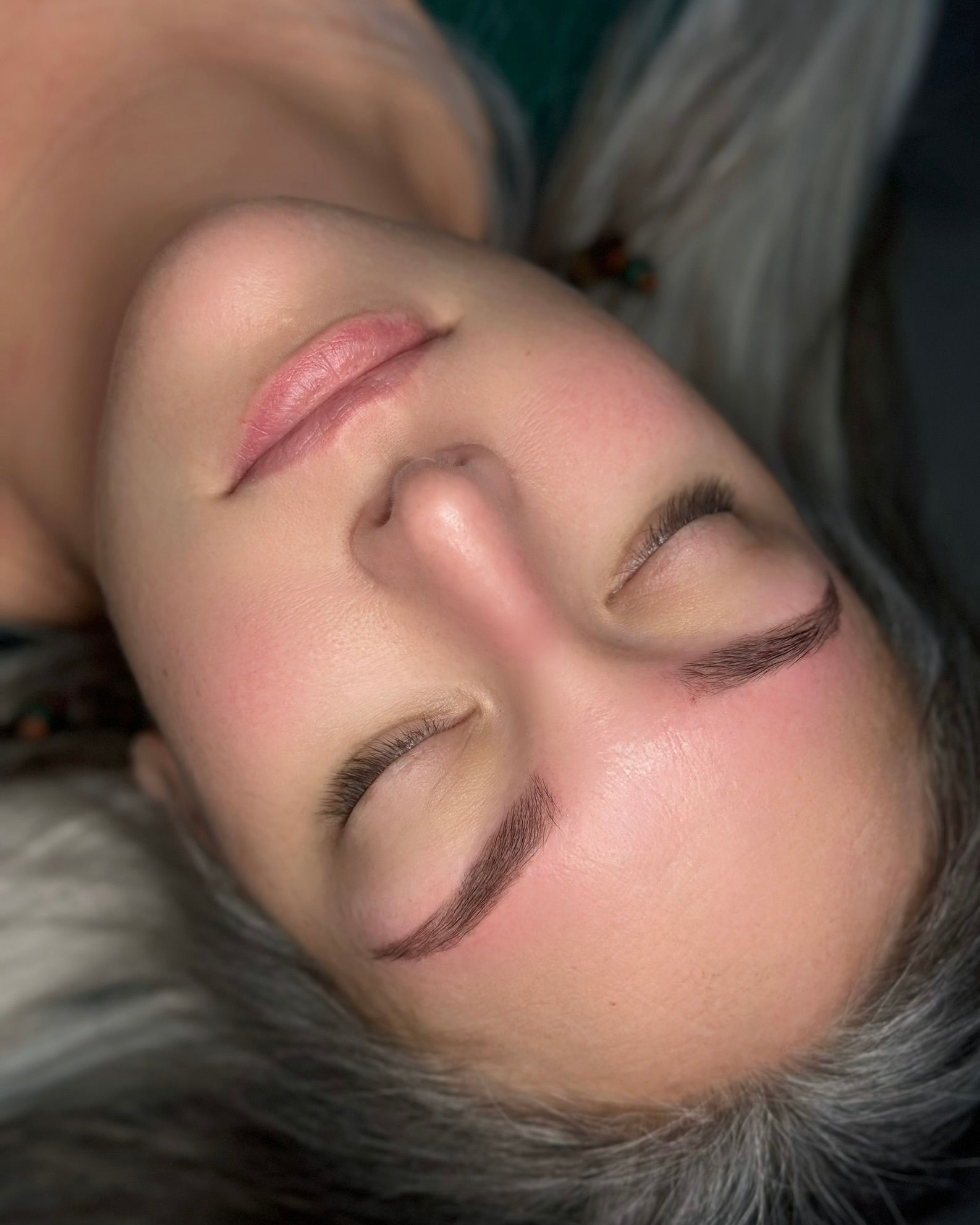 Woman with closed eyes, fresh eyebrow tattoo. Blush on cheeks, neutral skin tone, lying down.