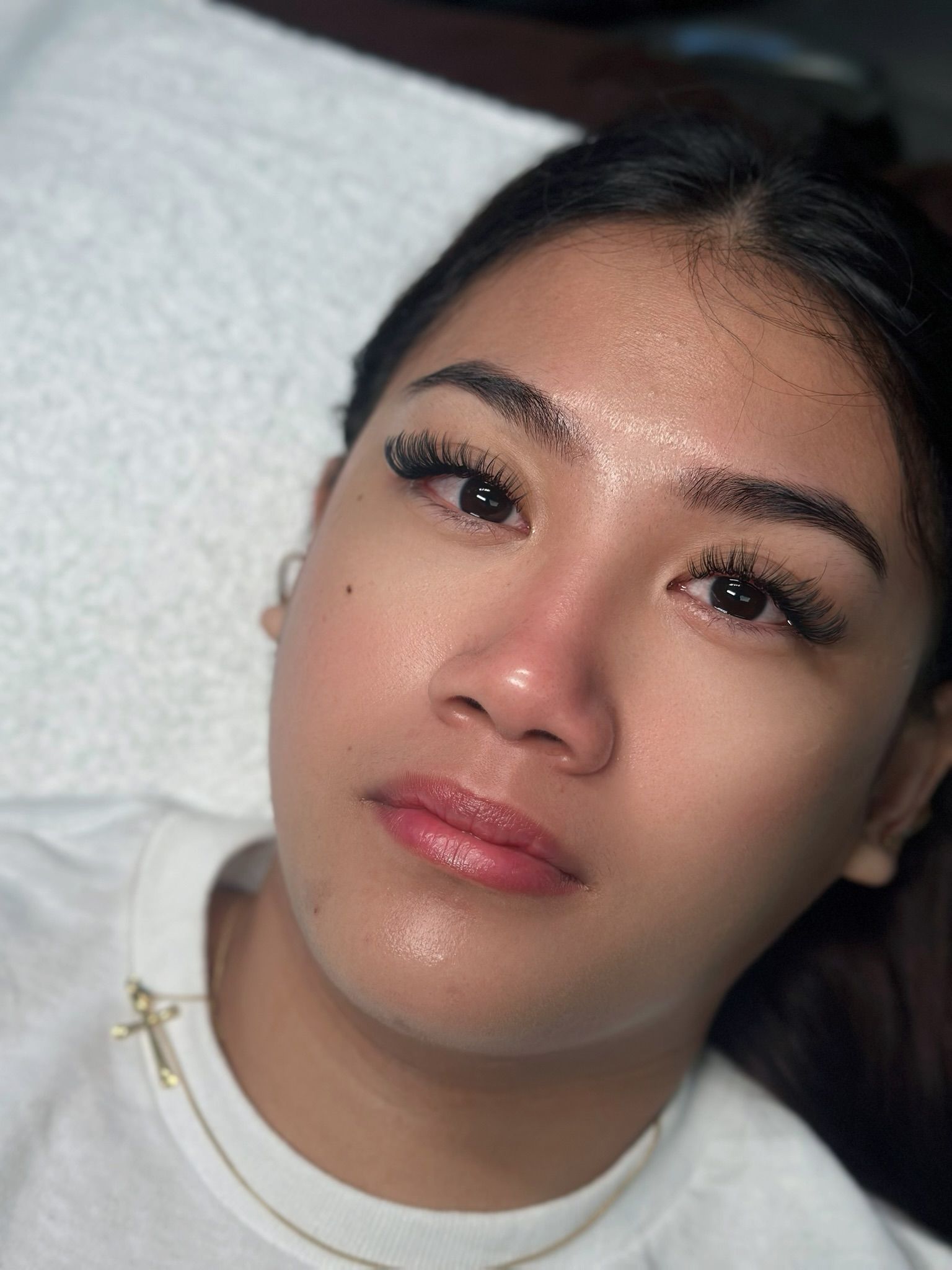 Woman with dark lashes and a gold necklace, lying on a white surface, looking at the camera.