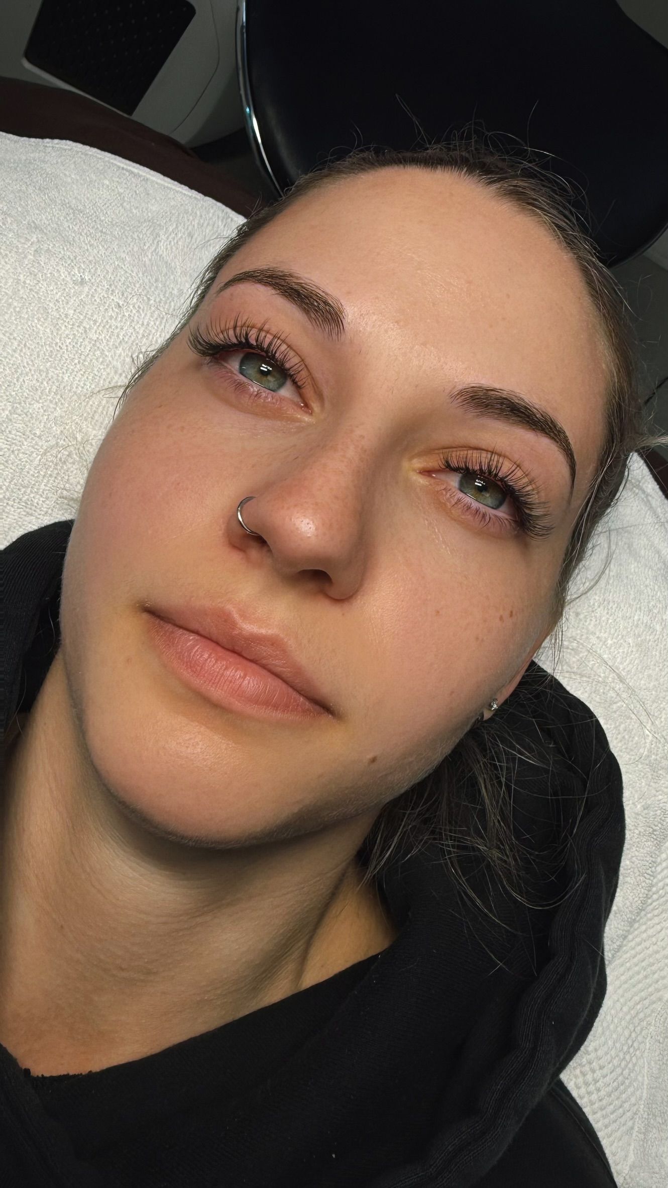 Woman lying on a white surface, freshly done eyebrows, nose ring, wearing a black top.