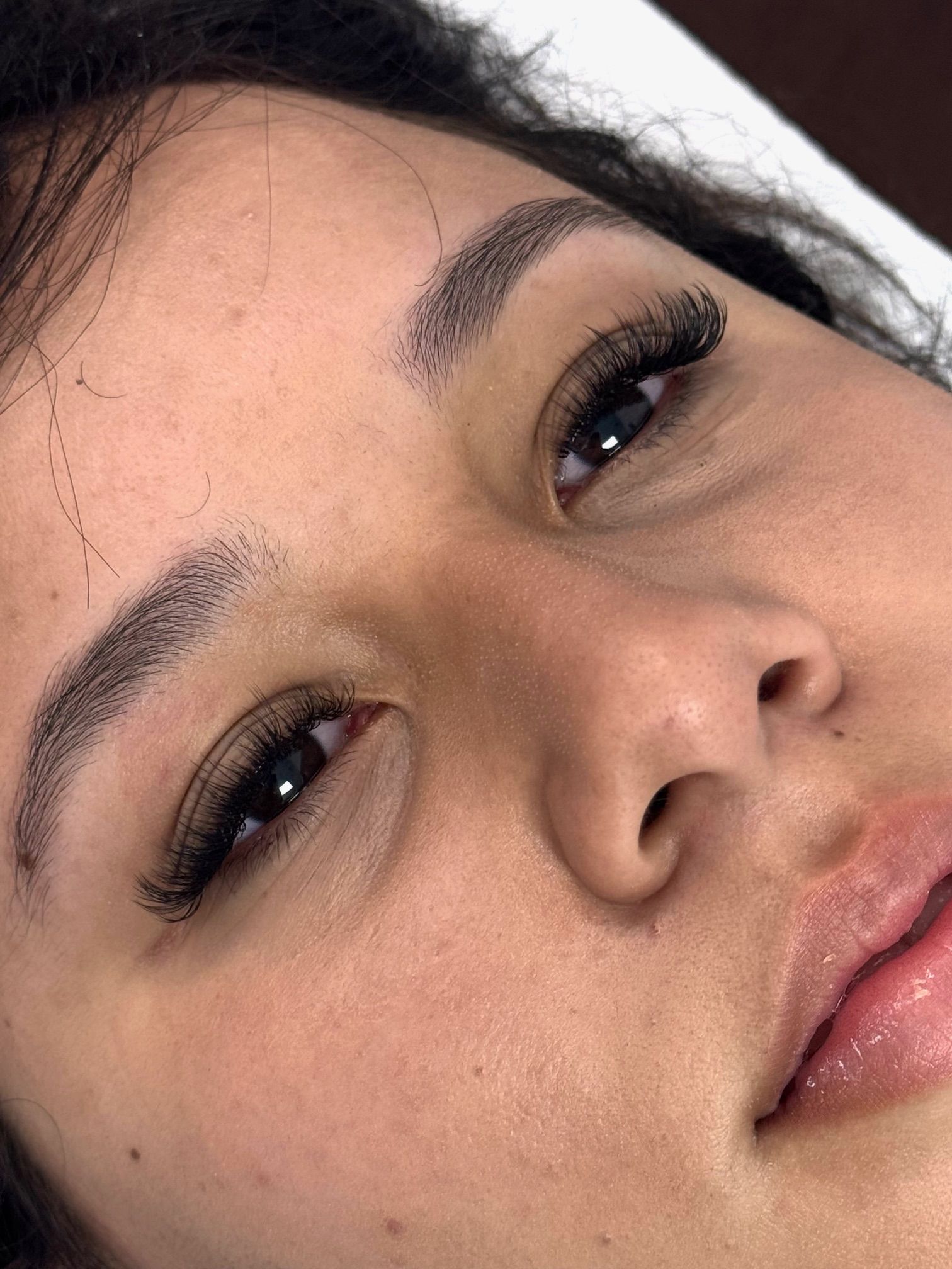 Close-up of a person's face with darkened eyebrows and eyelashes. Skin appears textured. Lips are glossy.