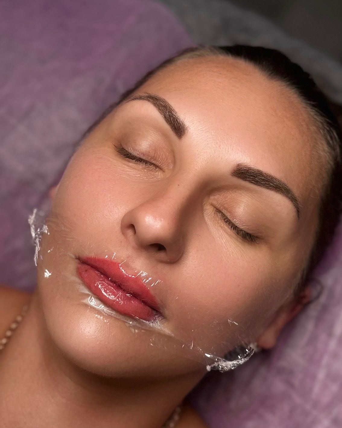Woman with closed eyes, lip gloss, and clear film on face, presumably after a facial treatment.