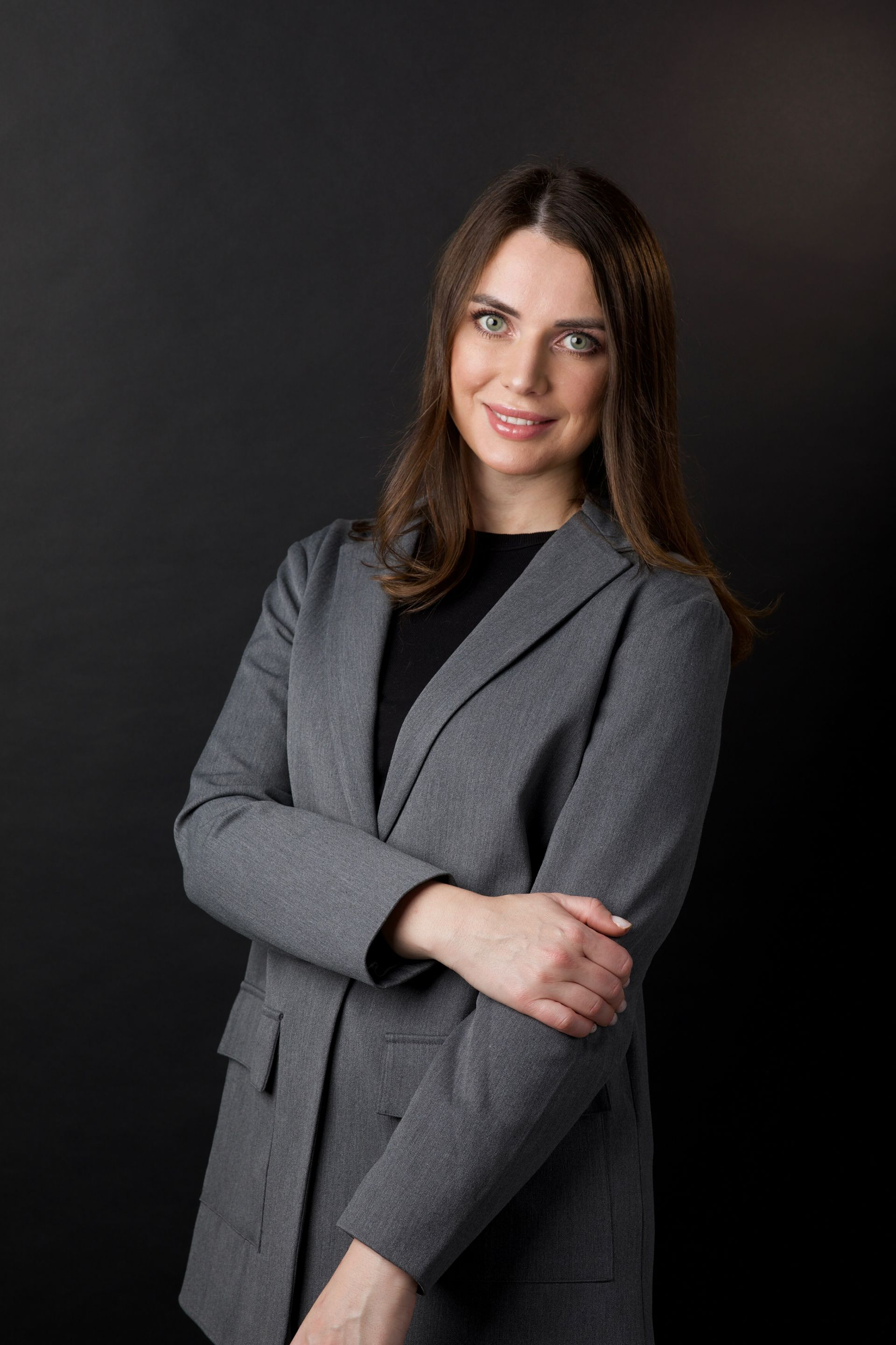 Woman in a gray blazer and black top smiles, arms crossed, set against a black background.