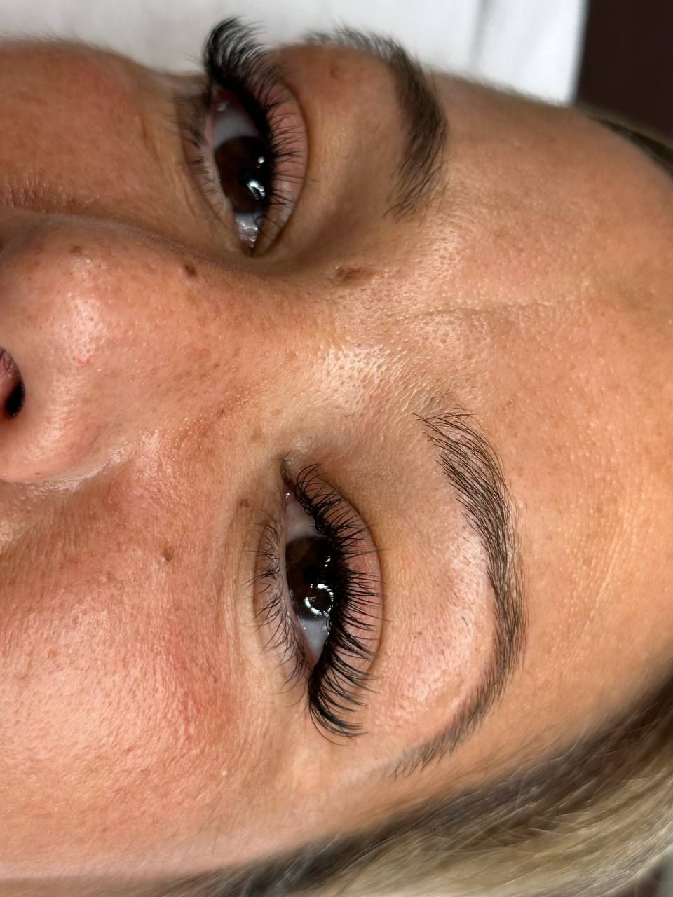 Close-up of a person's eyes and eyebrows, with dark eyelashes and tattooed eyebrows, skin is slightly red.