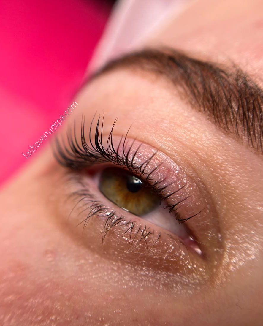Close-up of an eye with lifted lashes, light skin, and brown eyebrow. The eye is hazel and the background is pink.
