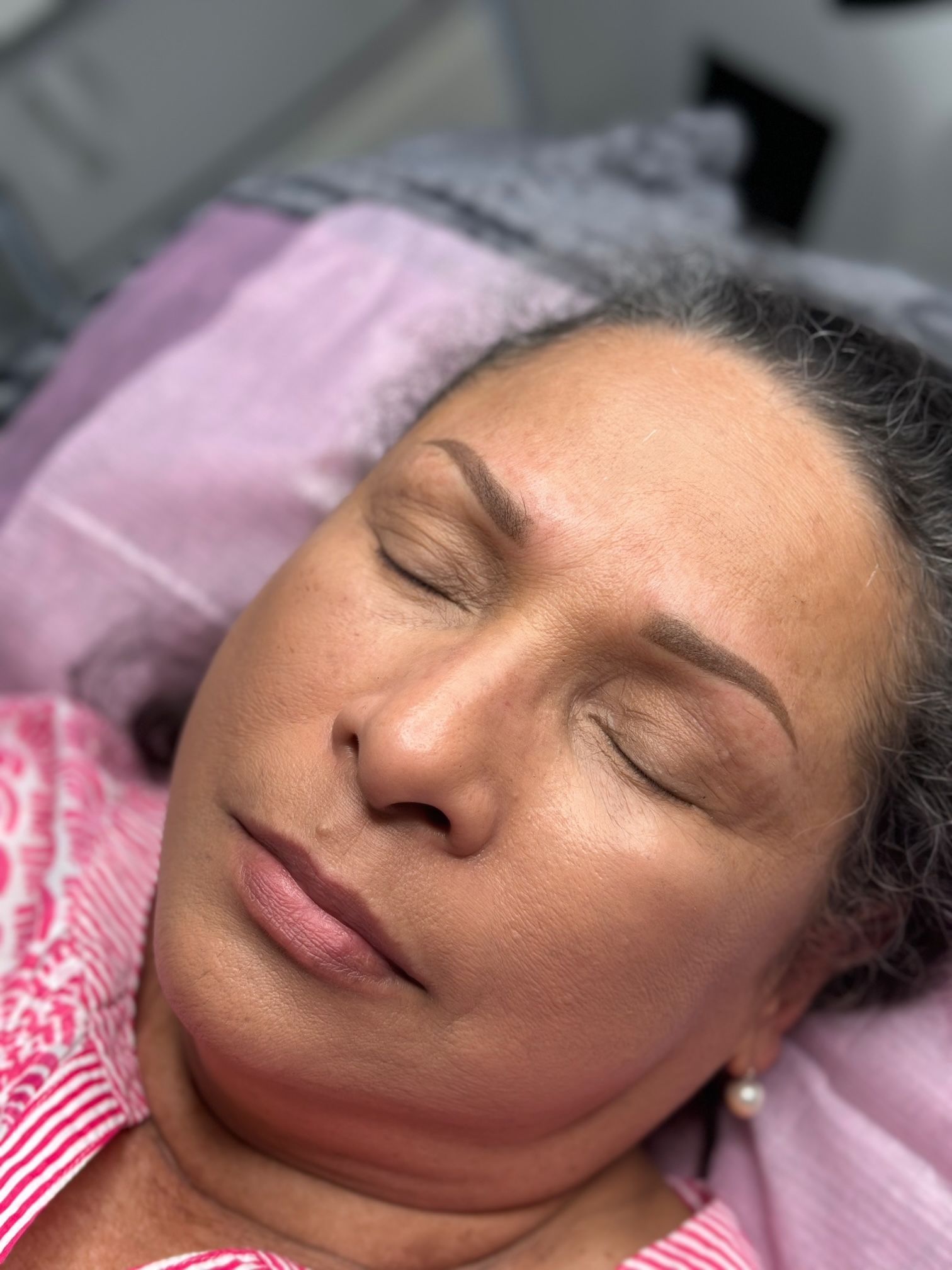 Woman with eyes closed, freshly done eyebrow tattoo, resting on a bed, light pink top.