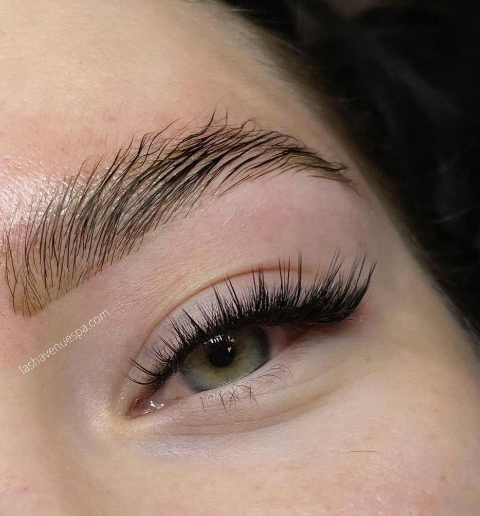 Close-up of eye with long lashes and brushed-up, dark eyebrows; skin is shiny.