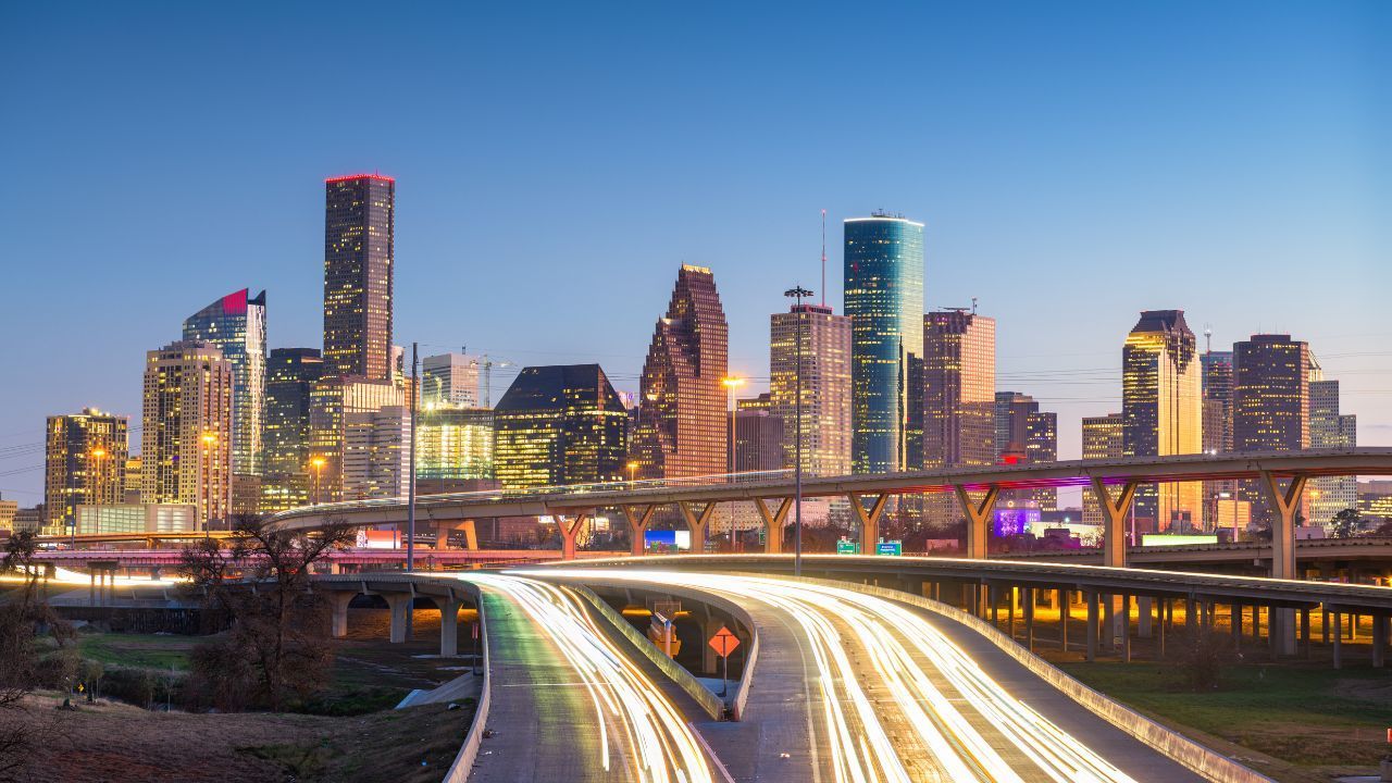 City skyline at dusk with illuminated towers and light trails from traffic on a highway.