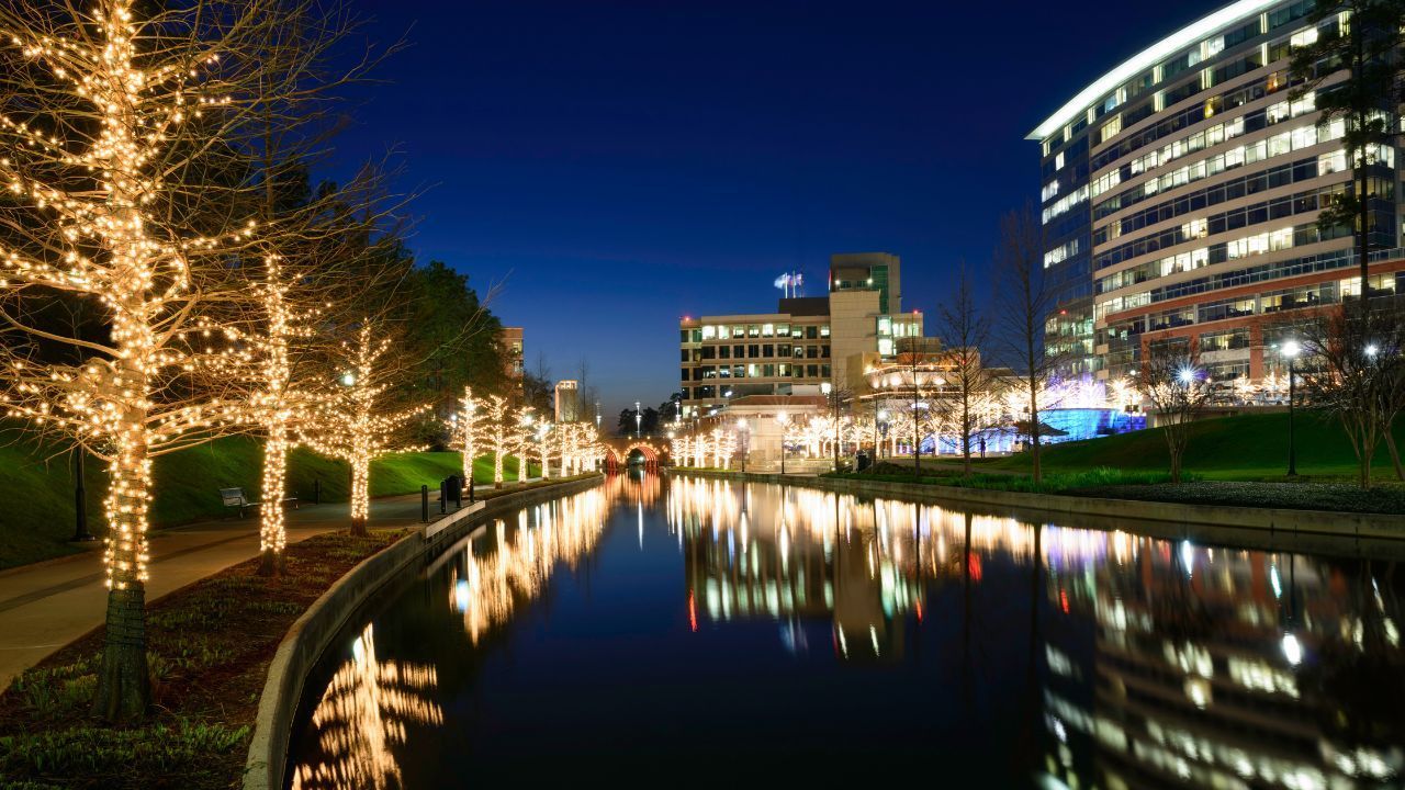 Night canal scene with lit trees, city buildings, and colorful reflections in the water.