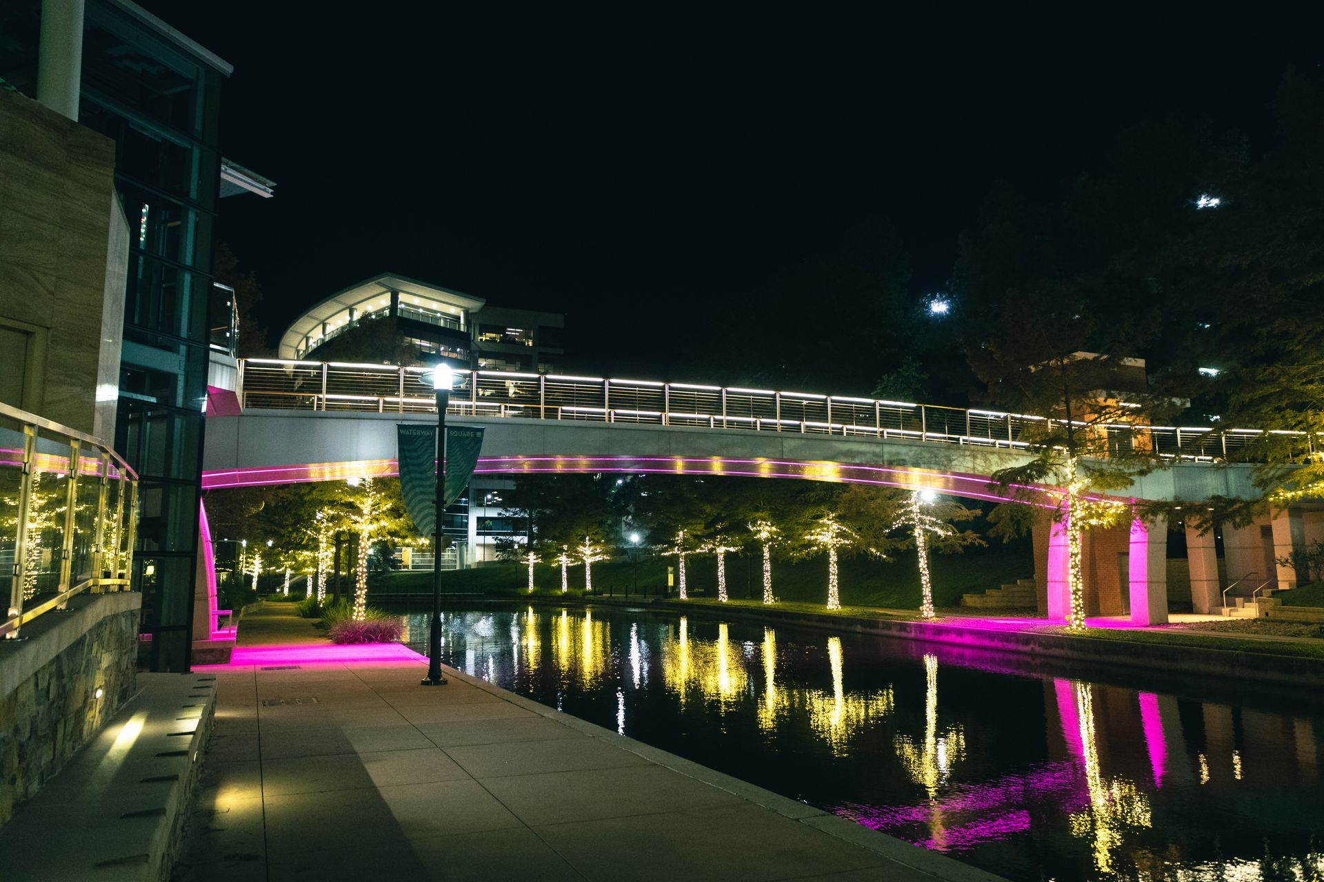 Nighttime illuminated pedestrian bridge over a canal, lit with pink and green lights.