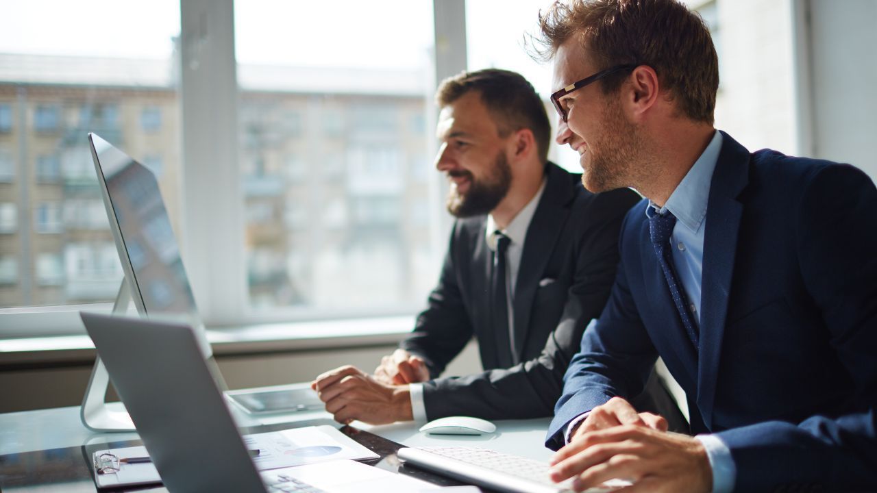 Two business professionals in suits work at a laptop in a bright office.
