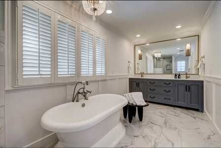 Bright modern bathroom with freestanding tub, gray vanity, large mirror, and white shutters