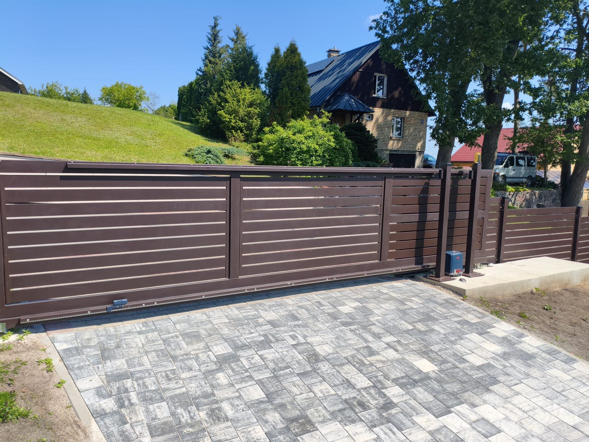 Brown metal gate and fence beside a paved driveway, with a house and trees on a sunny day.