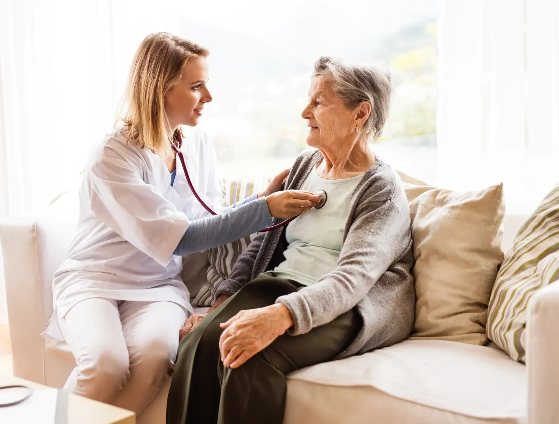 Nurse listens to a patient's heartbeat with a stethoscope. They are seated on a white sofa indoors  — Tewantin Medical Centre In Tewantin, QLD