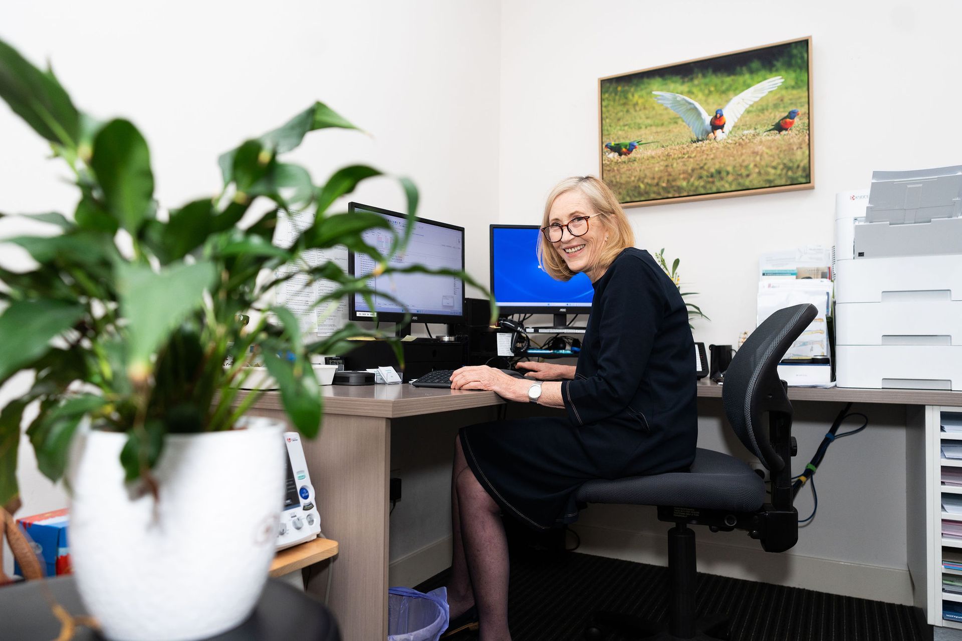 Woman sits at desk with two computer screens in office setting, smiling. Plant in foreground  — Tewantin Medical Centre In Tewantin, QLD