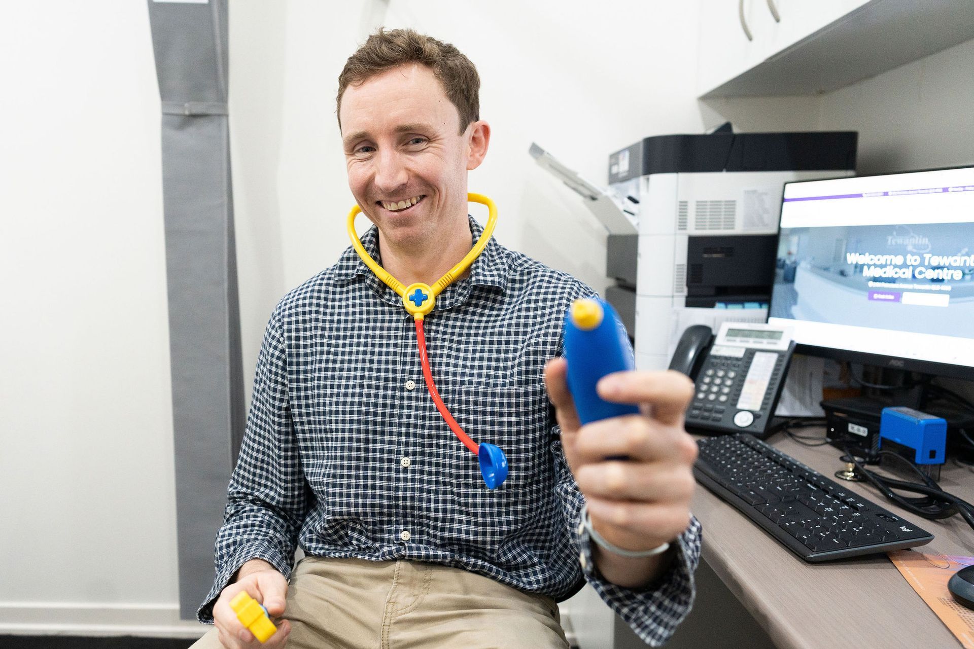 Doctor smiling, holding a toy stethoscope and medical instrument in an office setting  — Tewantin Medical Centre In Tewantin, QLD