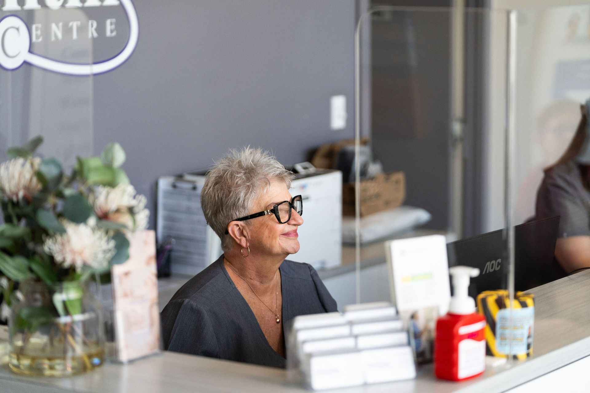Receptionist smiling behind a clear barrier at a medical center — Tewantin Medical Centre In Tewantin, QLD