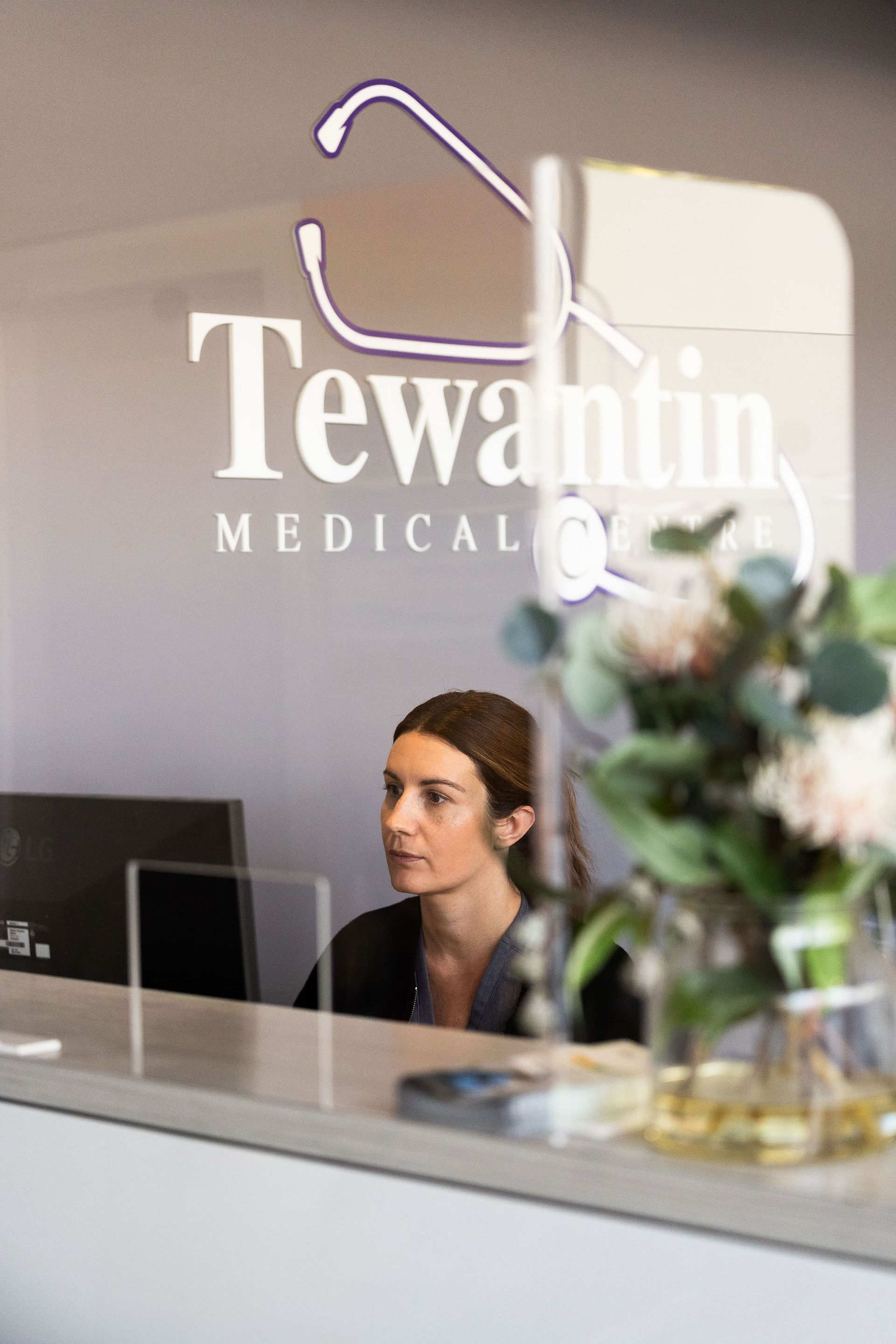 Receptionist at Tewantin Medical Center behind a desk with computer, clear barrier, and flower arrangement.