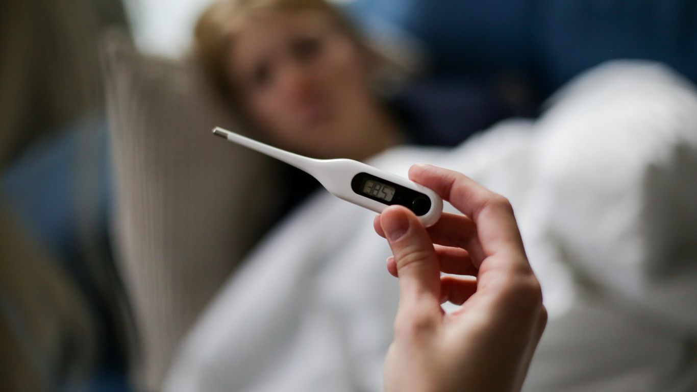 Person lying in bed while another hand holds a digital thermometer displaying a temperature of 38.5°C, indicating a fever.