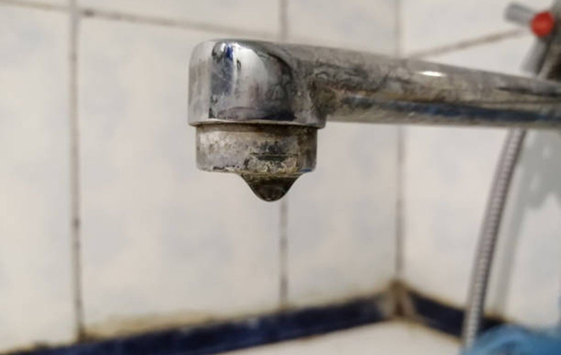 A dripping, dirty faucet with mineral buildup against tiled wall.