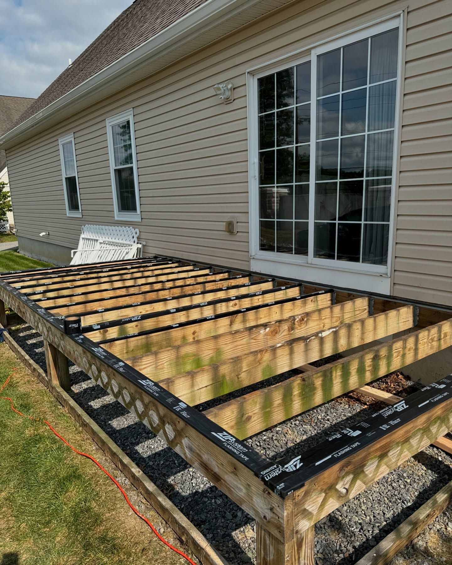 Partially constructed deck frame attached to a house with sliding glass doors.  Green grass and gravel are visible.