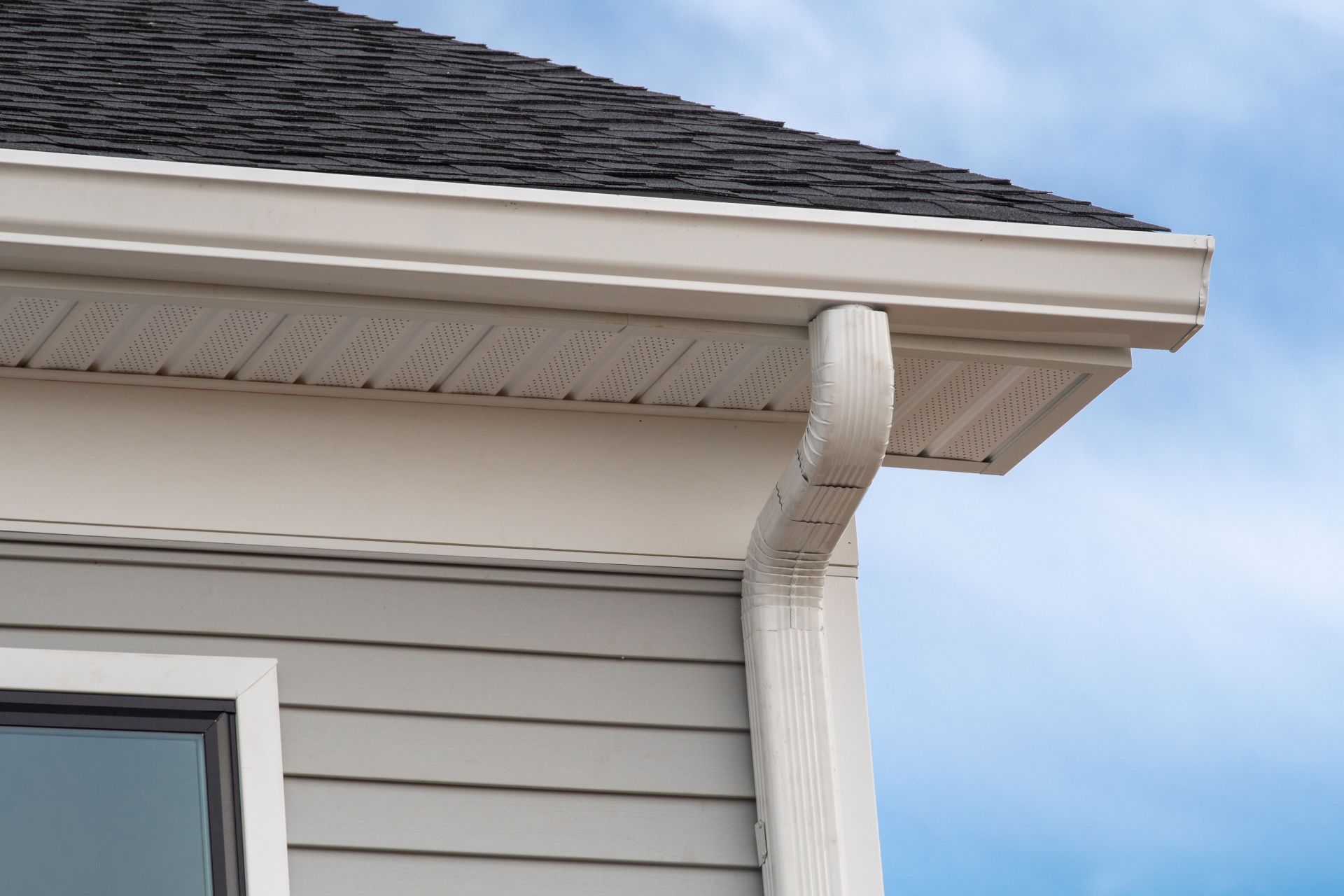 Beige gutters and downspout on a house with gray siding and a dark roof, against a blue sky.