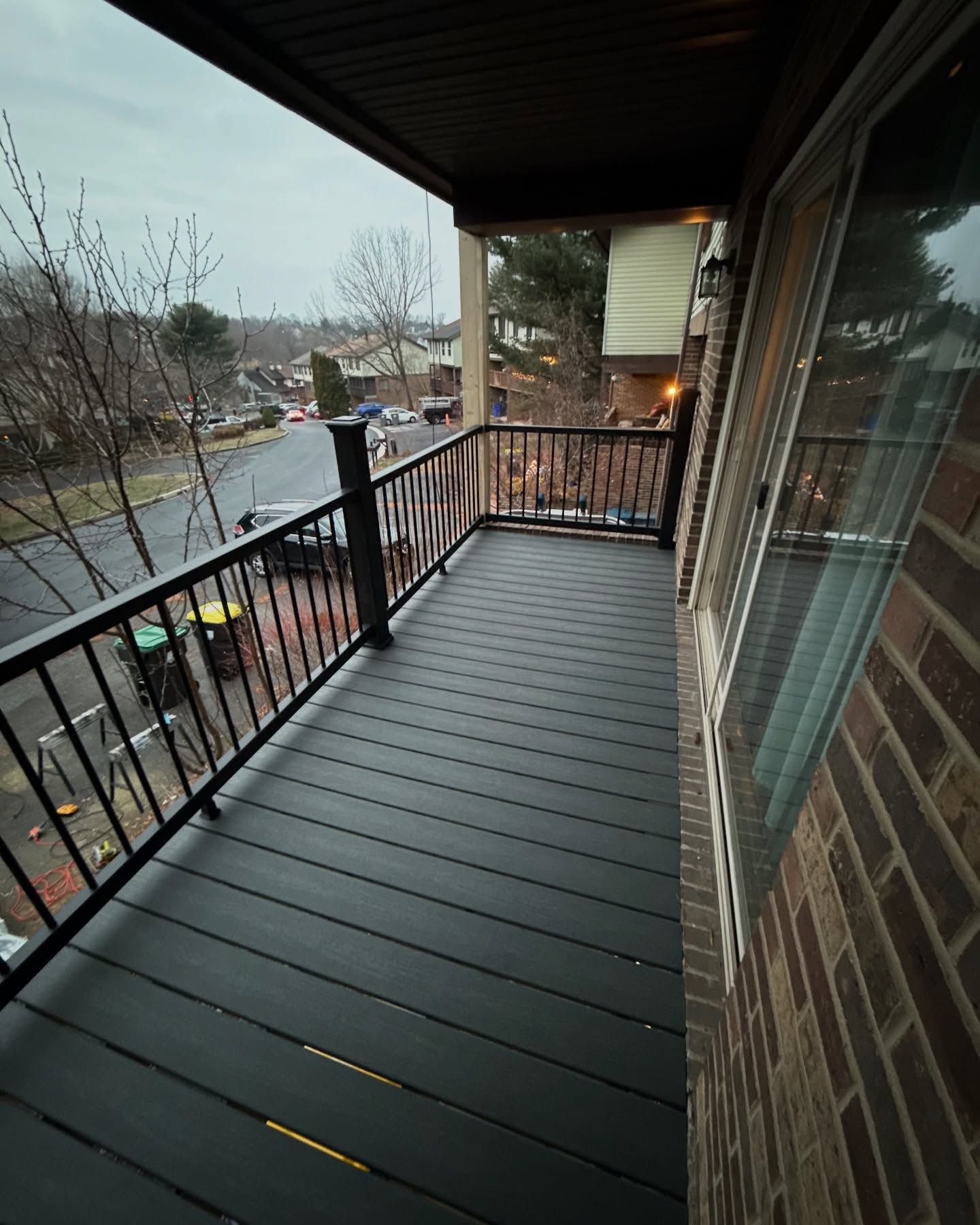 Balcony with black flooring and railing, overlooking a street with trees and overcast sky.