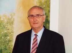 A man in a dark suit, light shirt, and striped tie stands in front of a painted landscape wall mural.