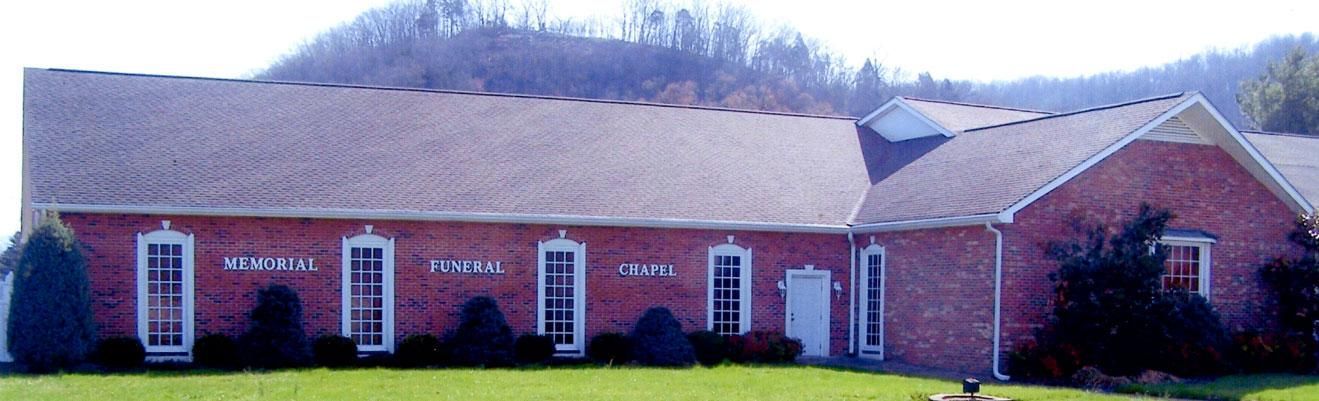 A brick building with a gabled roof, white window trim, and a small white door, set against a backdrop of green hills.