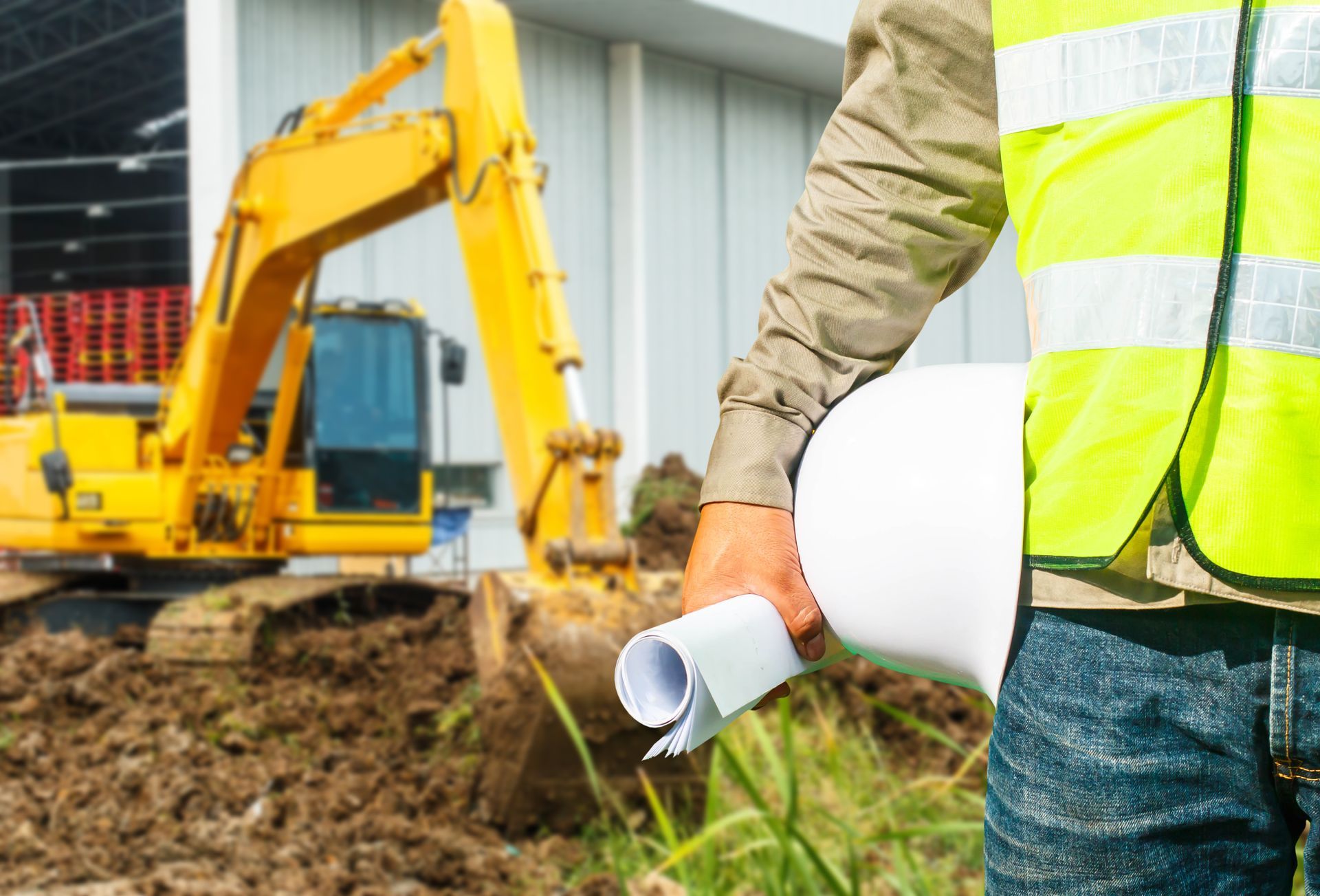 A construction worker is holding a helmet and a blueprint at a construction site.