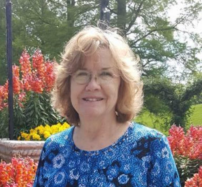 A woman wearing glasses and a blue shirt is standing in front of flowers.