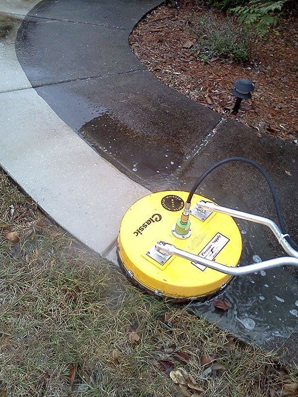 A yellow circular pressure washer surface cleaner is being used to clean a concrete walkway.
