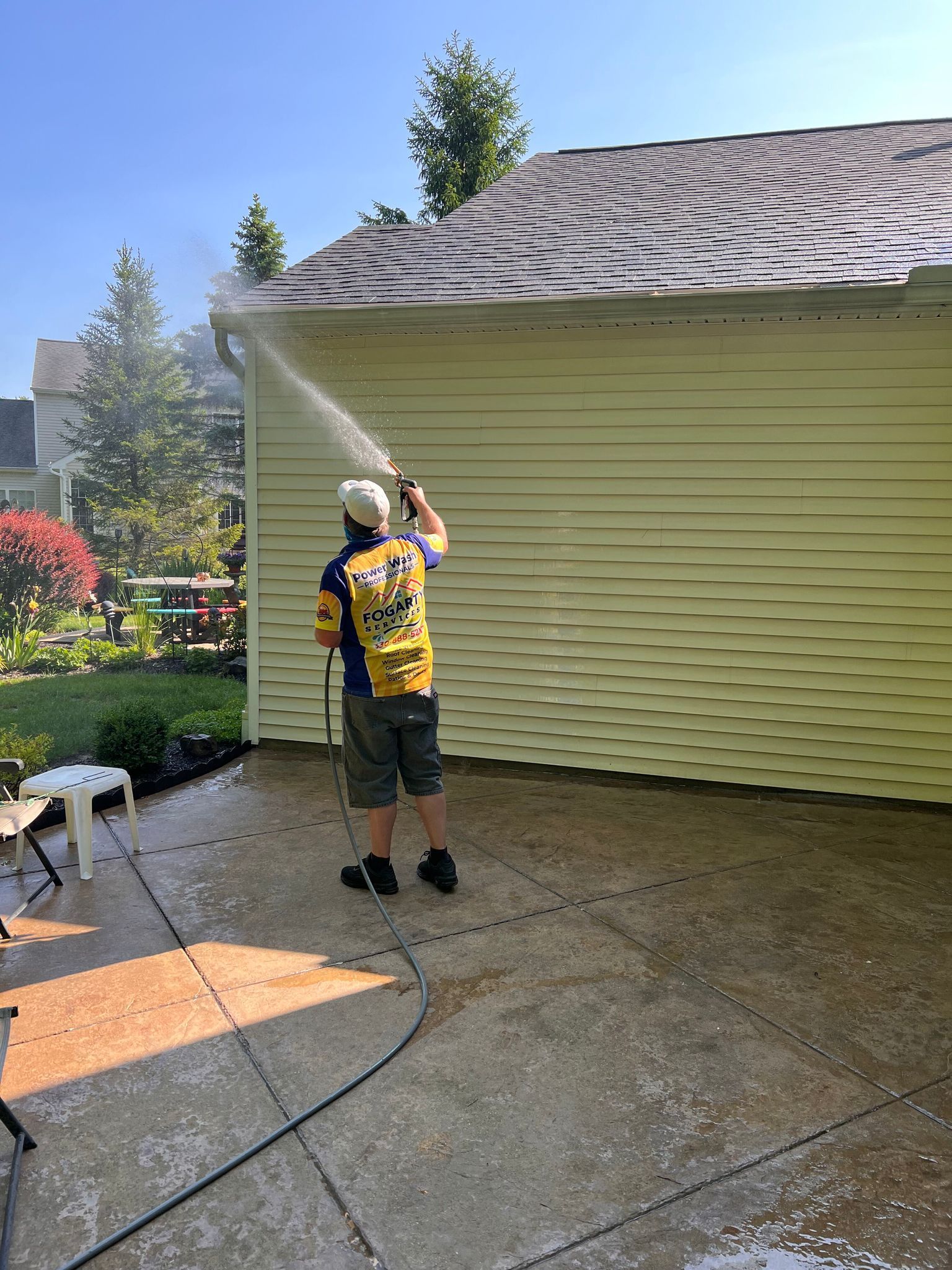 A person wearing a bright yellow backpack sprays water on the light yellow siding of a house in a residential yard.