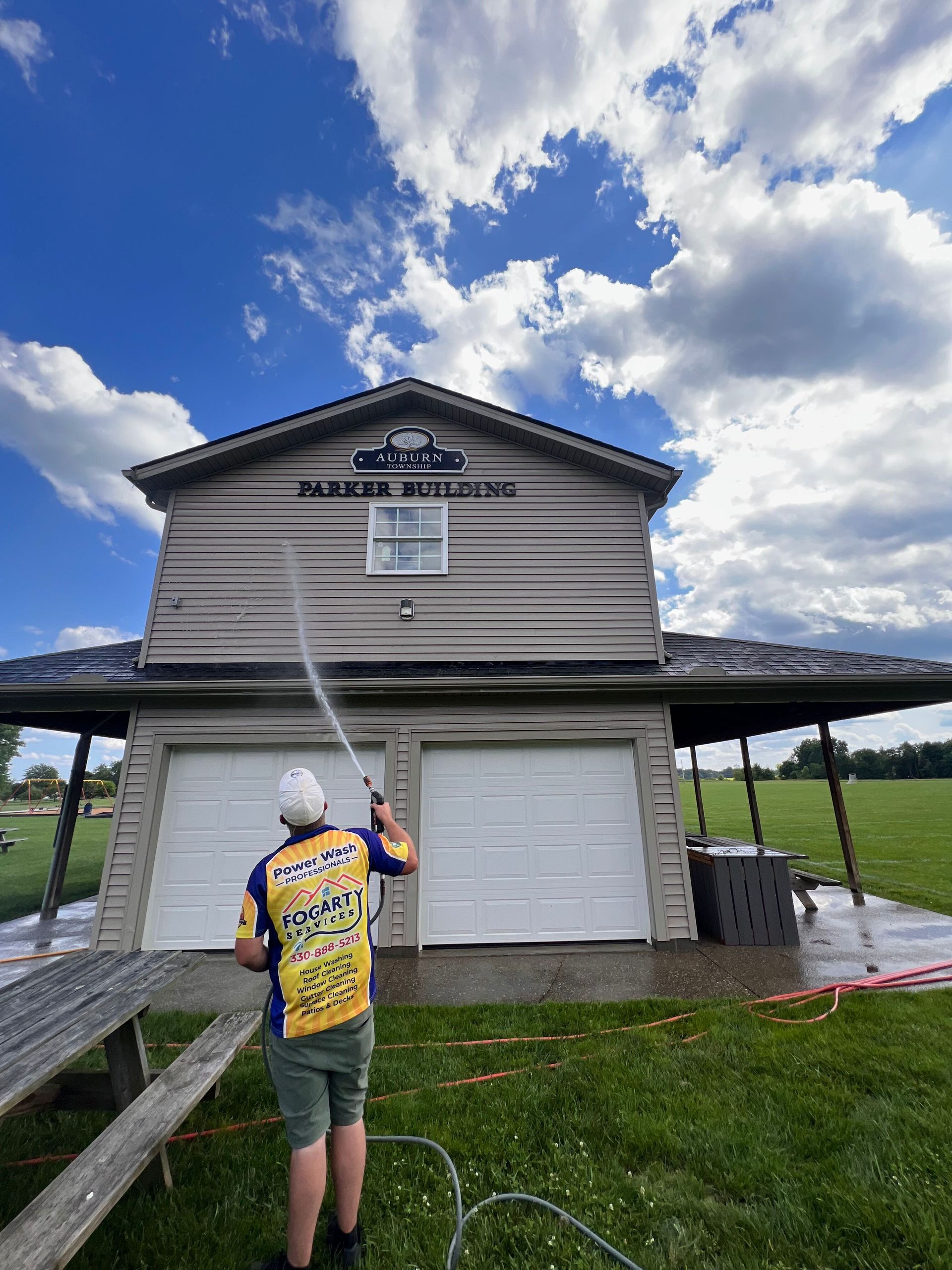 A person sprays water from a hose onto a two-story building with white garage doors and a sunny, cloudy sky overhead.
