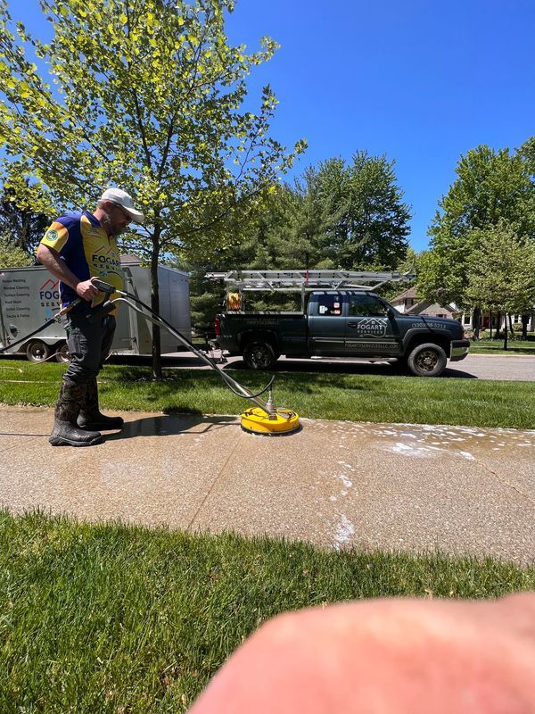 A worker uses a circular surface cleaner to power wash a concrete driveway on a sunny day near a parked utility truck.