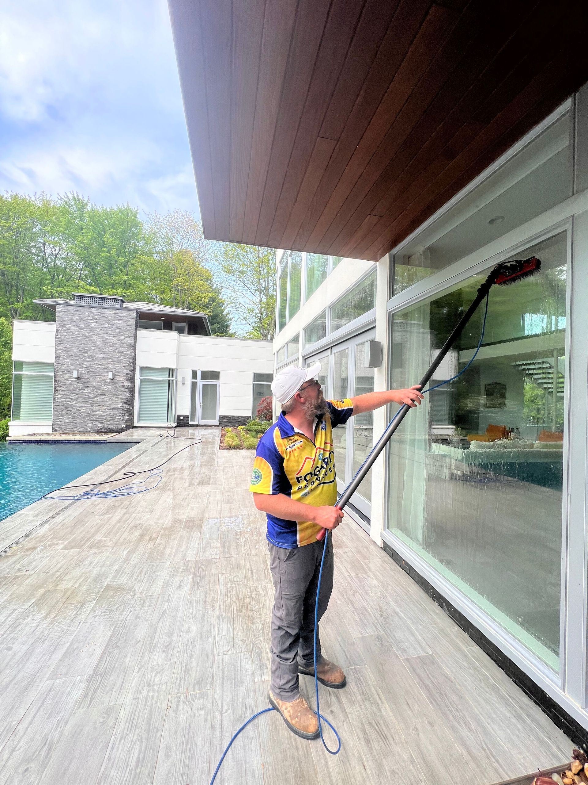 A person using an extended pole brush to clean the windows of a modern home with a patio and pool.