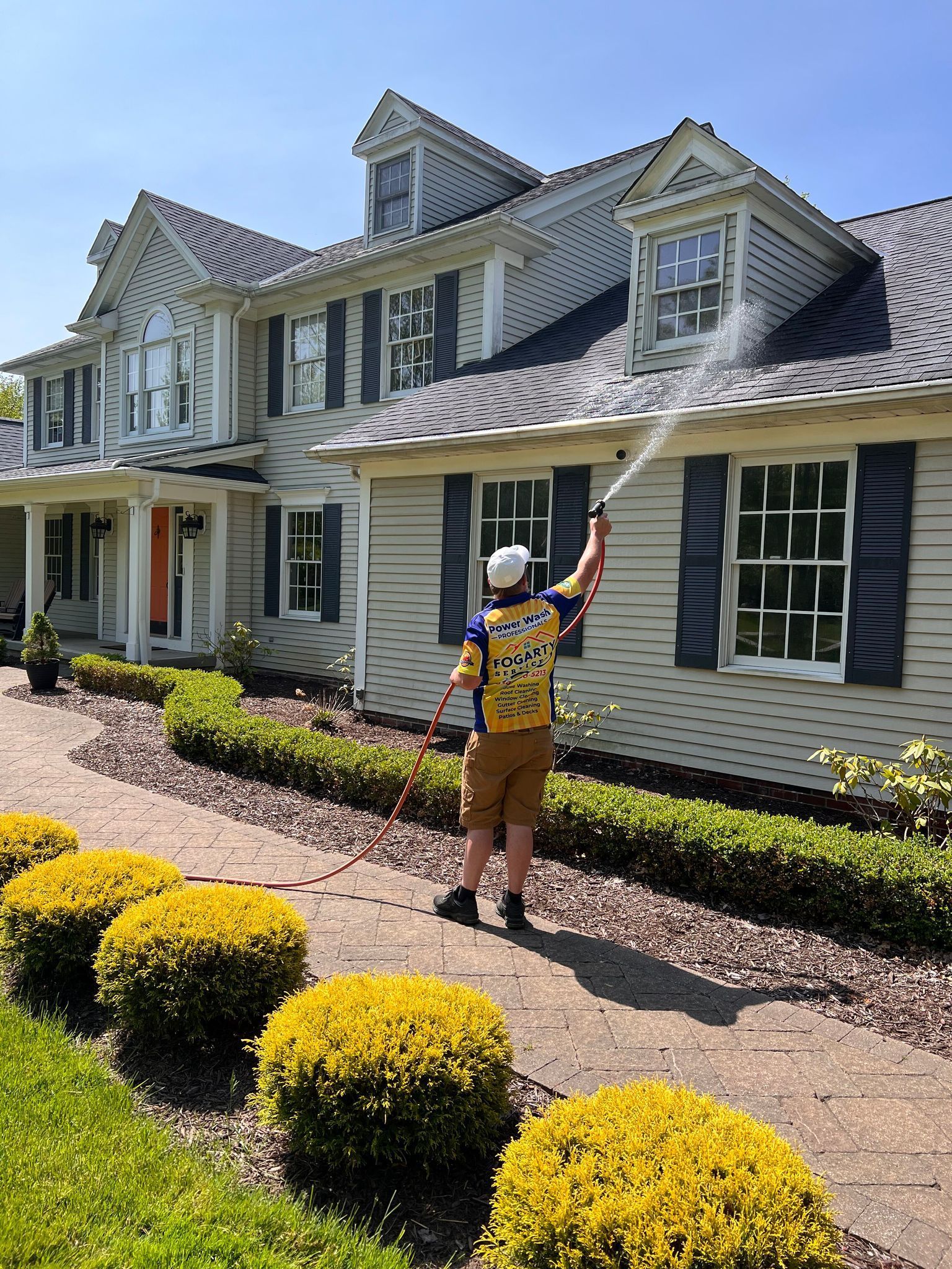 A worker wearing a yellow and blue shirt power washes the roof of a large house with a manicured front garden.
