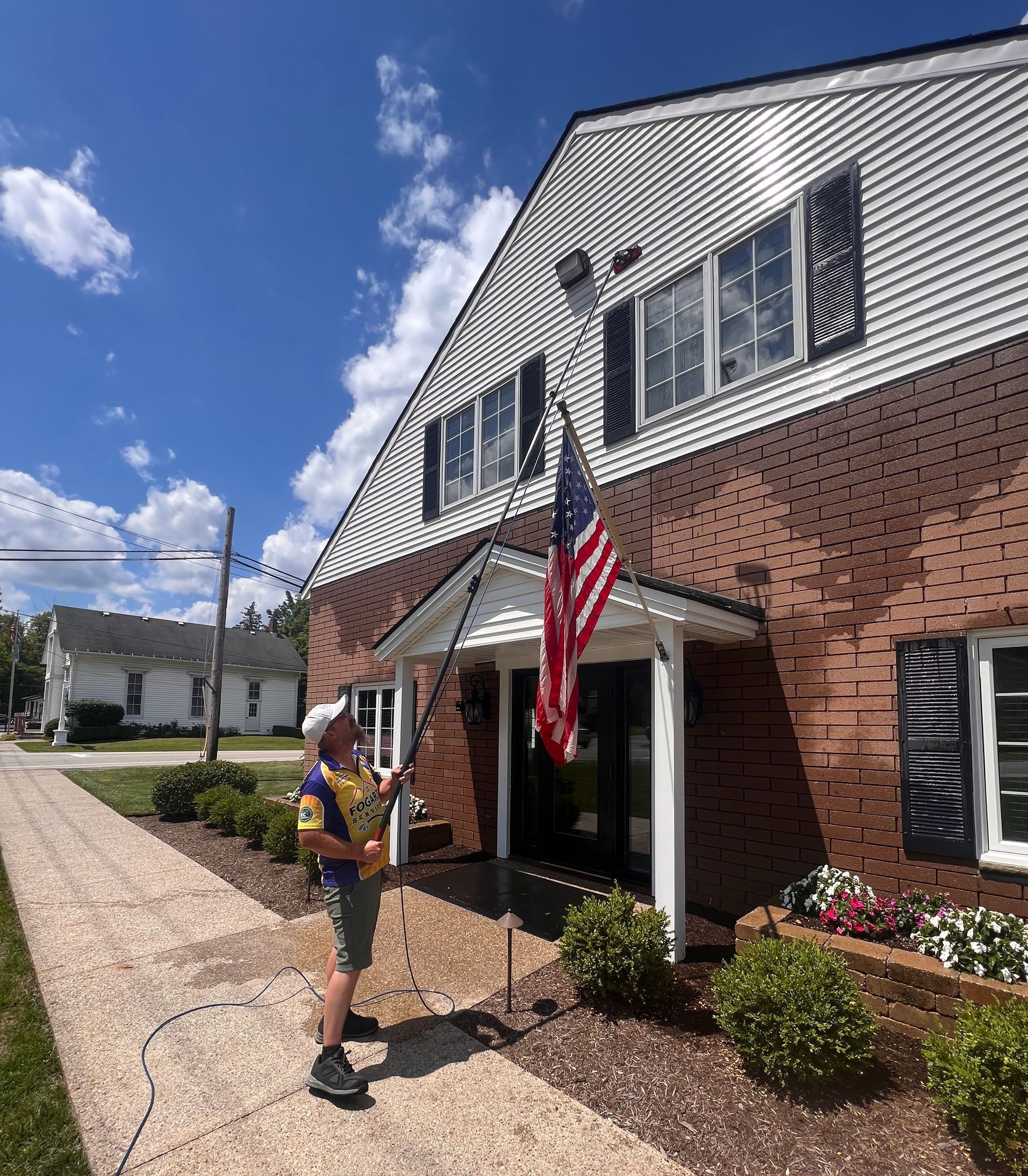 A person in a yellow and blue shirt stands on a sidewalk, using a long pole to adjust an American flag on a brick building.