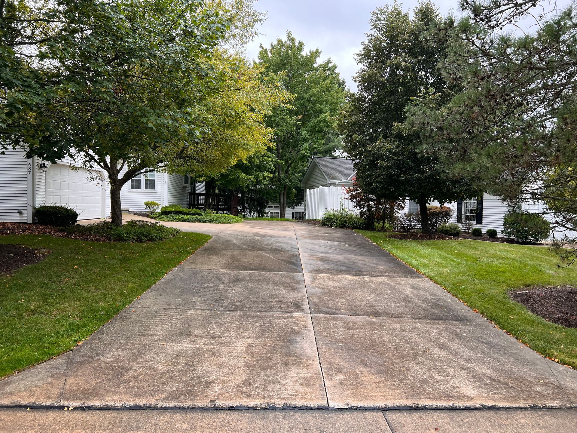 A concrete driveway leads toward houses surrounded by green trees under a cloudy sky.