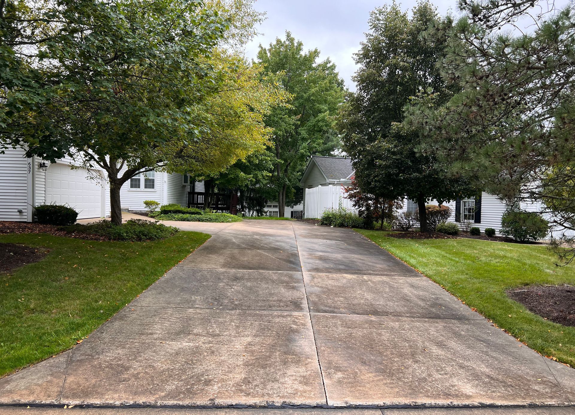 A weathered concrete driveway leads to a white house surrounded by lush green trees and manicured lawns under a cloudy sky.
