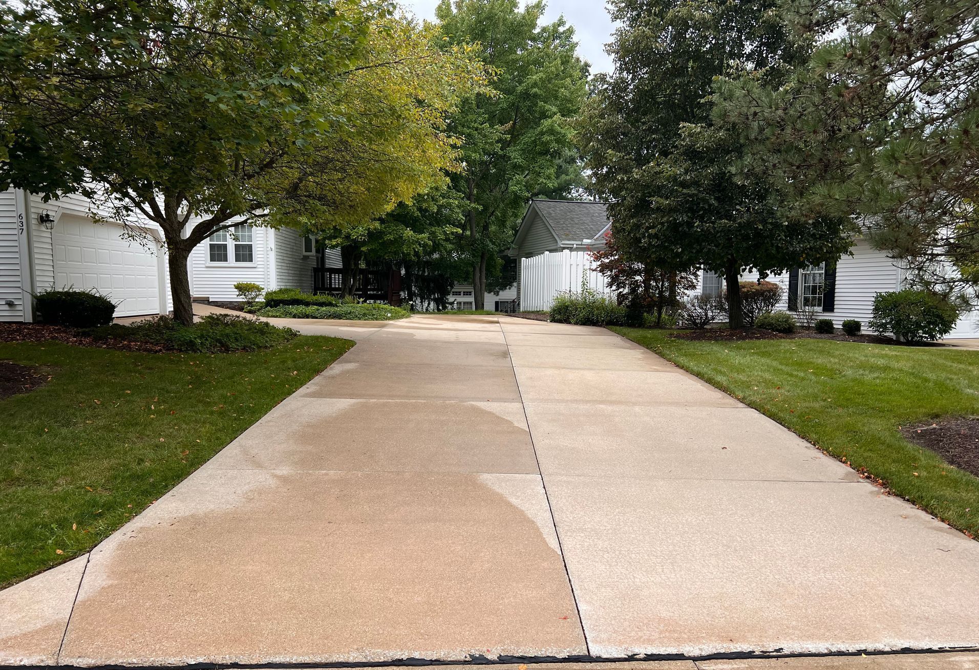 A paved driveway leads toward two white houses surrounded by green trees and lawns under a cloudy sky.