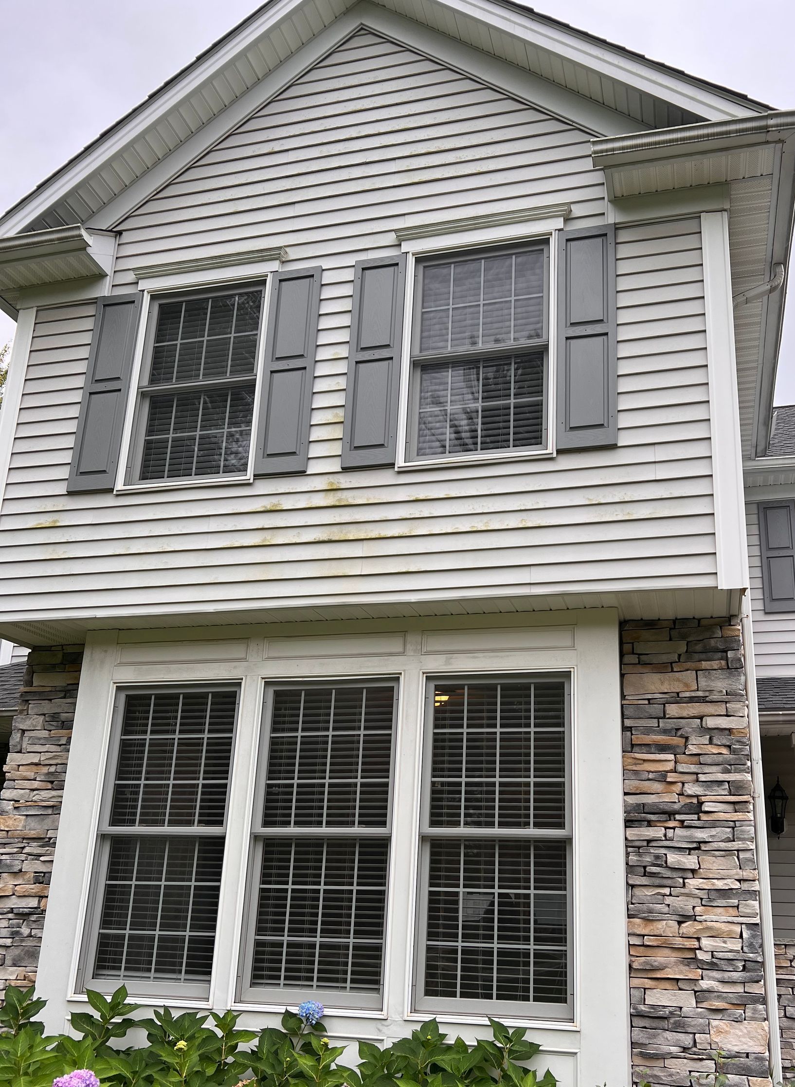 A two-story house exterior featuring horizontal white siding, a stone-veneer base, and gray-trimmed windows with shutters.