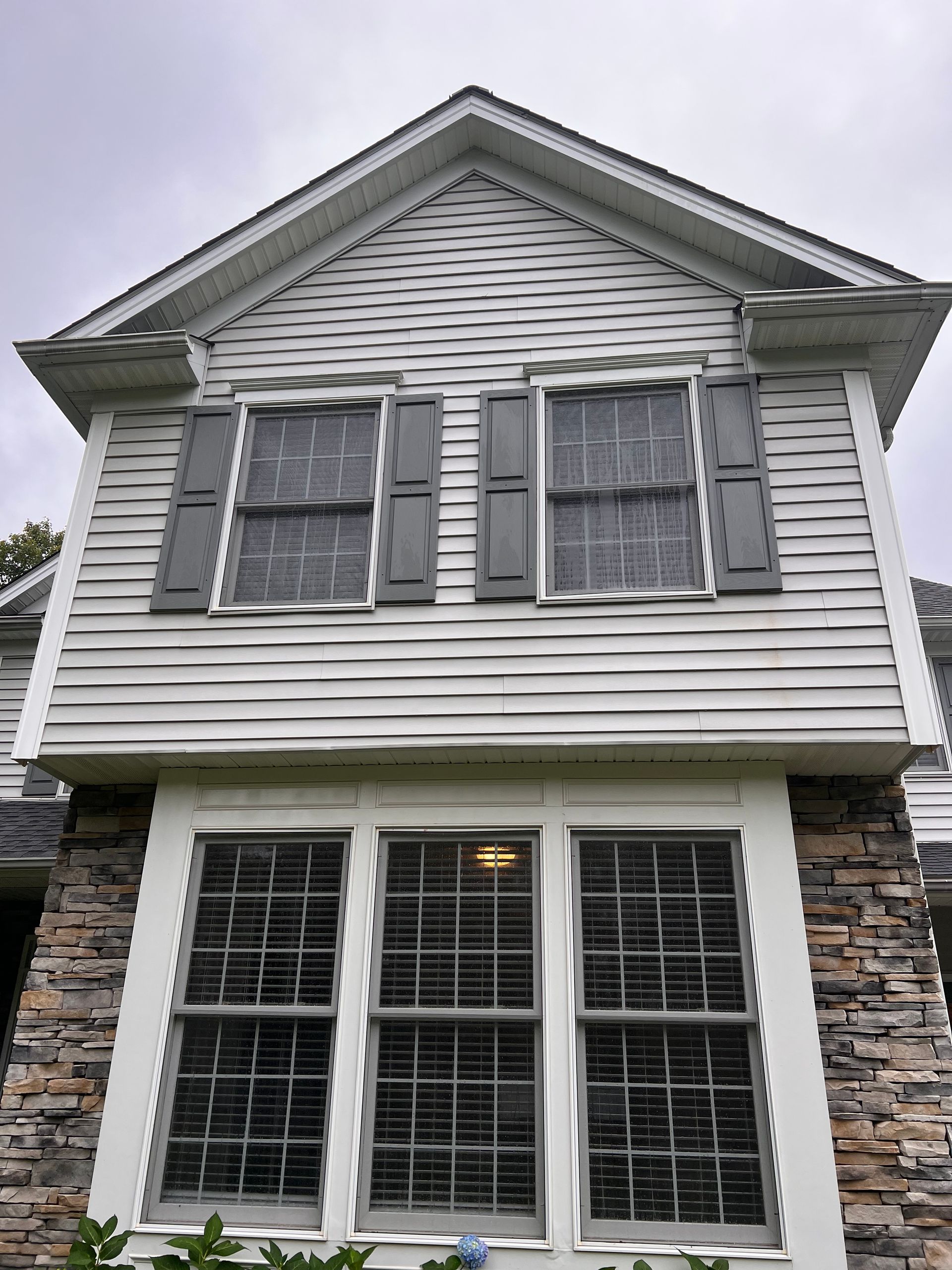 A two-story house exterior featuring light horizontal siding, dark grey shutters, and a lower stone-facade bay window.