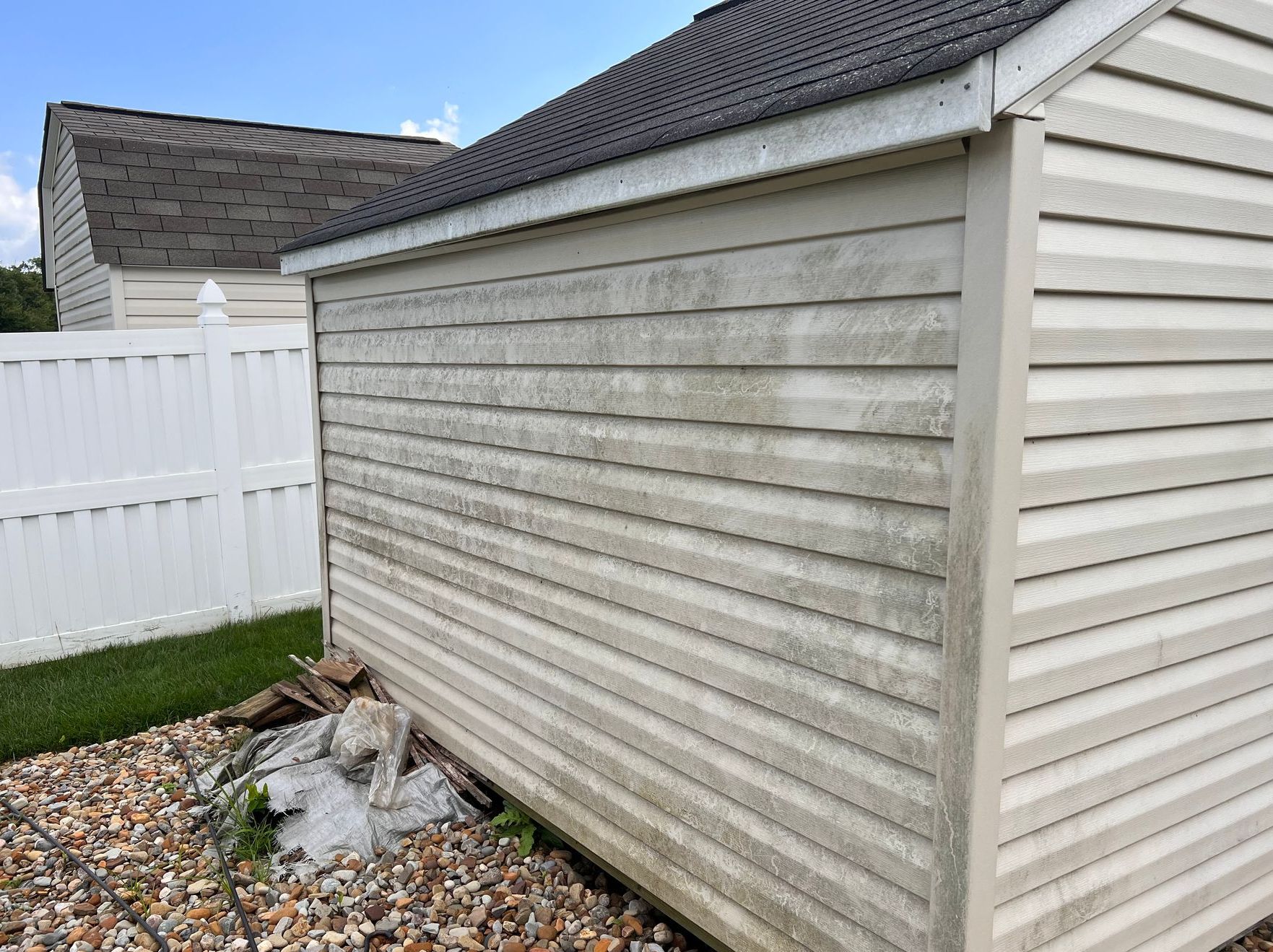 An exterior view of a cream-colored storage shed with dirty, stained siding, standing next to a white privacy fence.