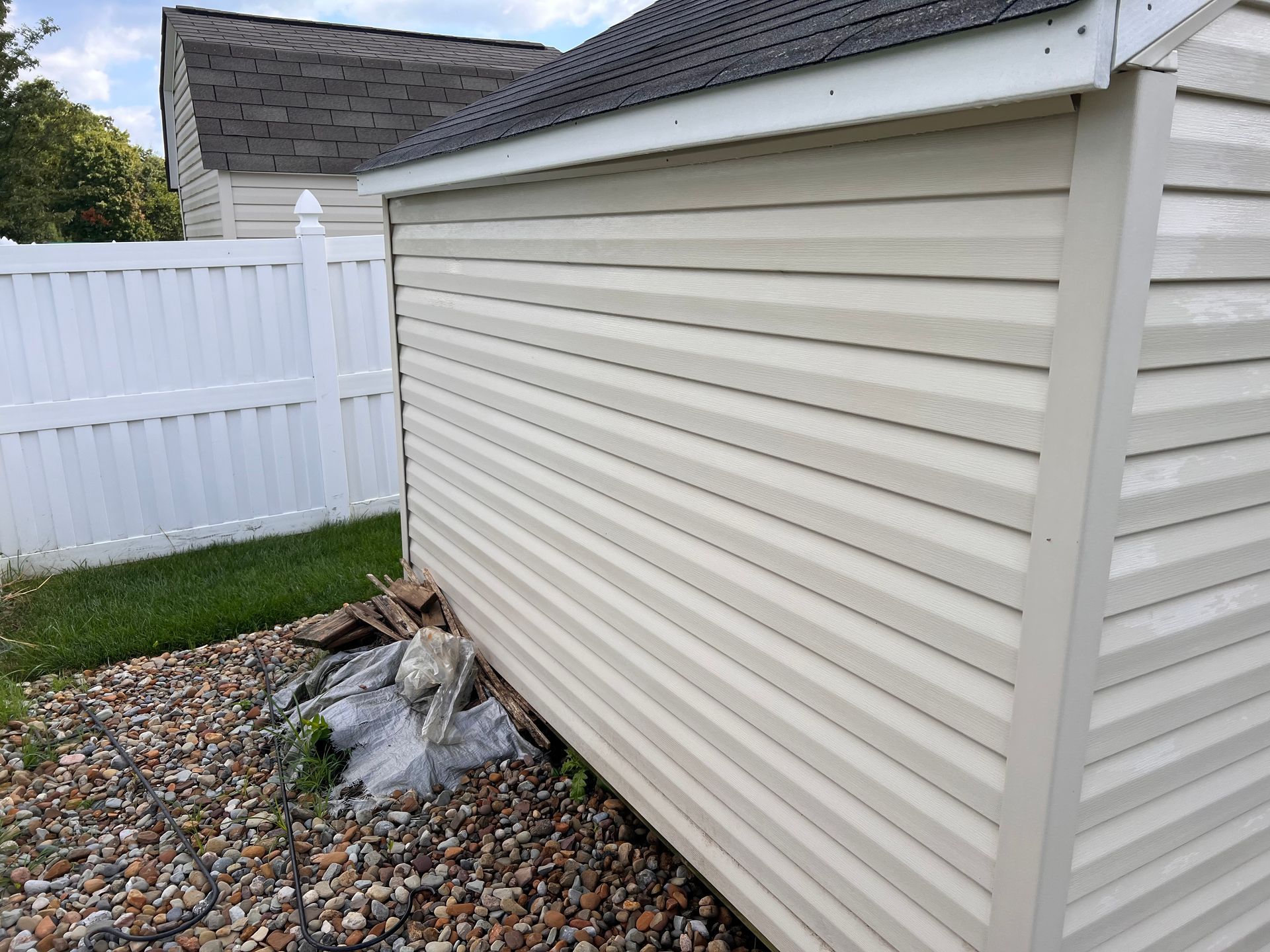 A beige vinyl-sided shed sits next to a white privacy fence, with a small pile of rocks and debris at its corner.