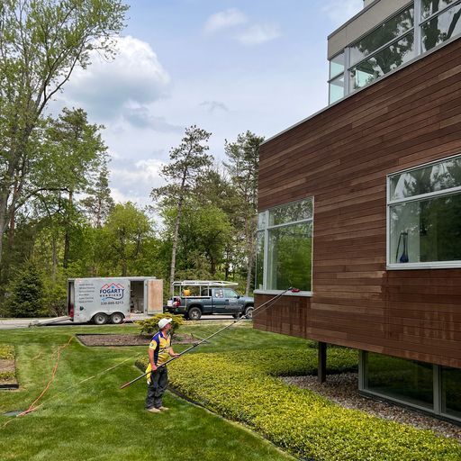 A person uses a long water-fed pole to clean the exterior windows of a modern, wood-sided building on a sunny day.