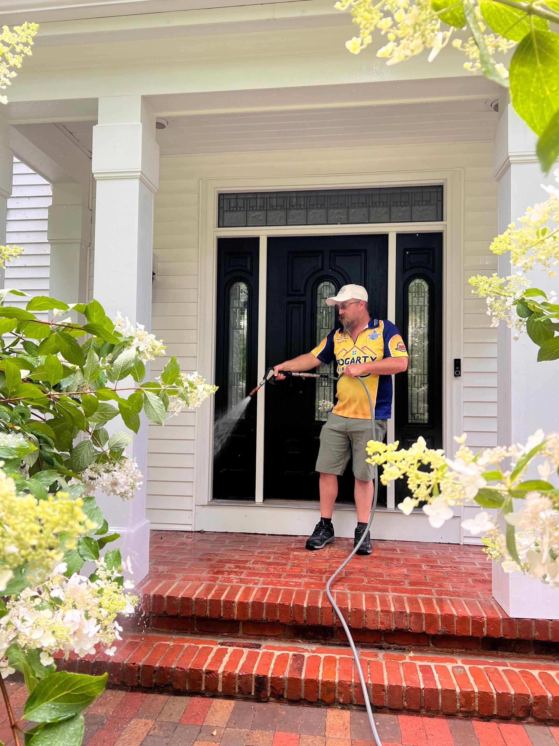 A person in a yellow and blue shirt uses a pressure washer to clean a dark front door on a brick porch.