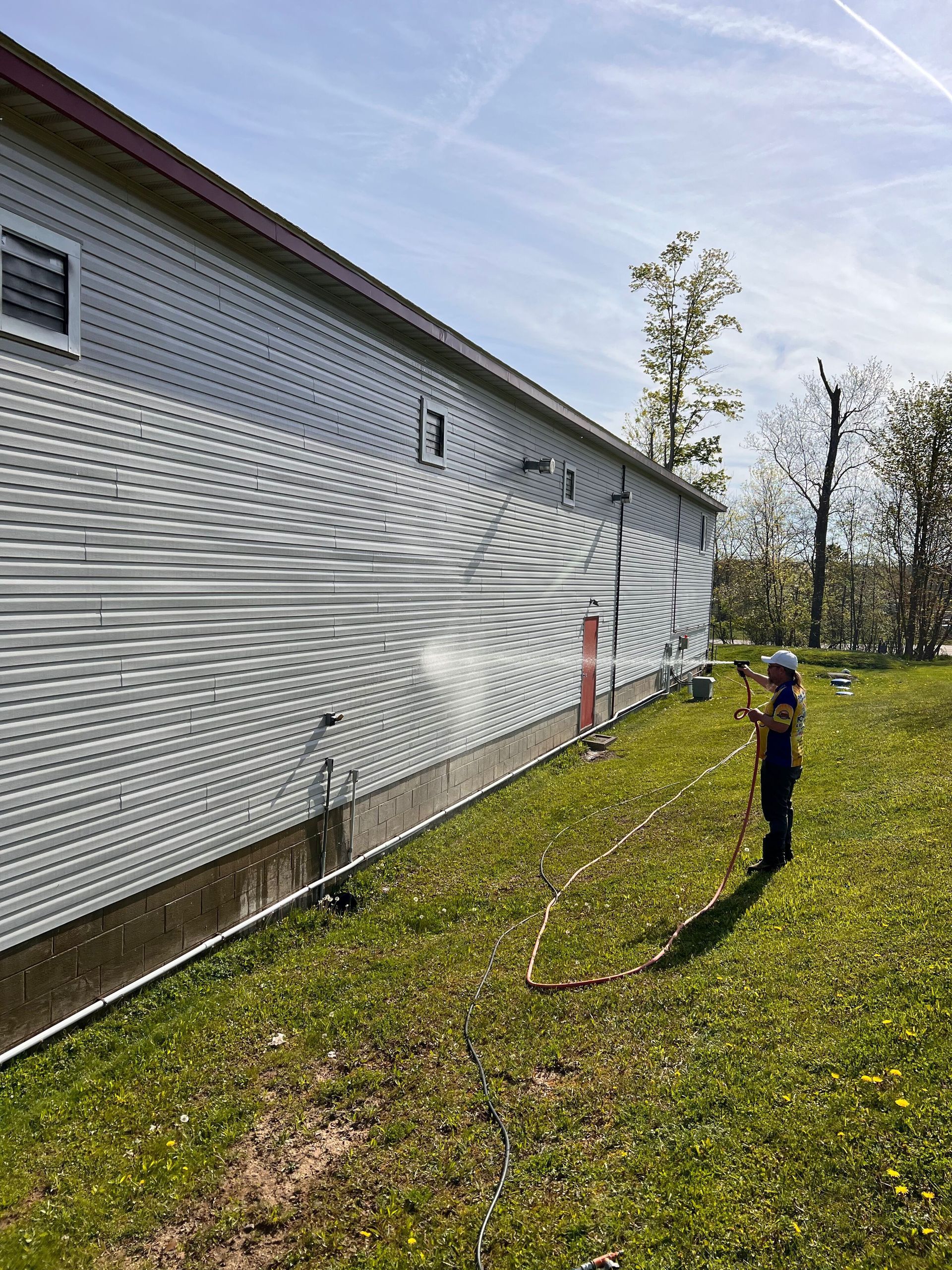 A person wearing a hard hat sprays water from a hose onto the side of a long, gray building on a sunny, grassy day.