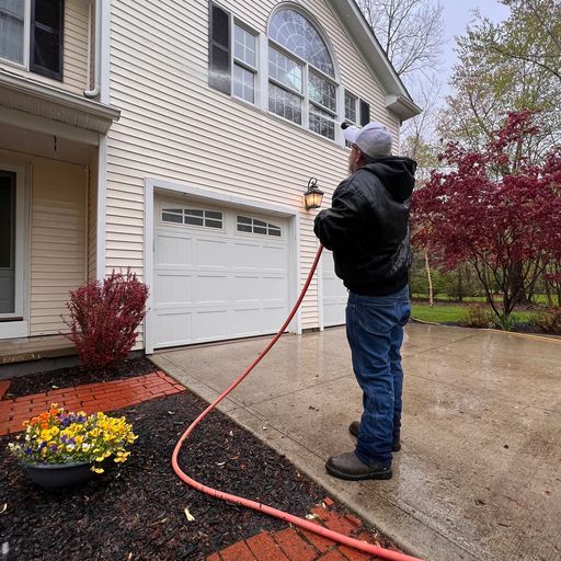 A person in a black jacket and white cap uses a power washer to clean the light-colored siding of a suburban house.