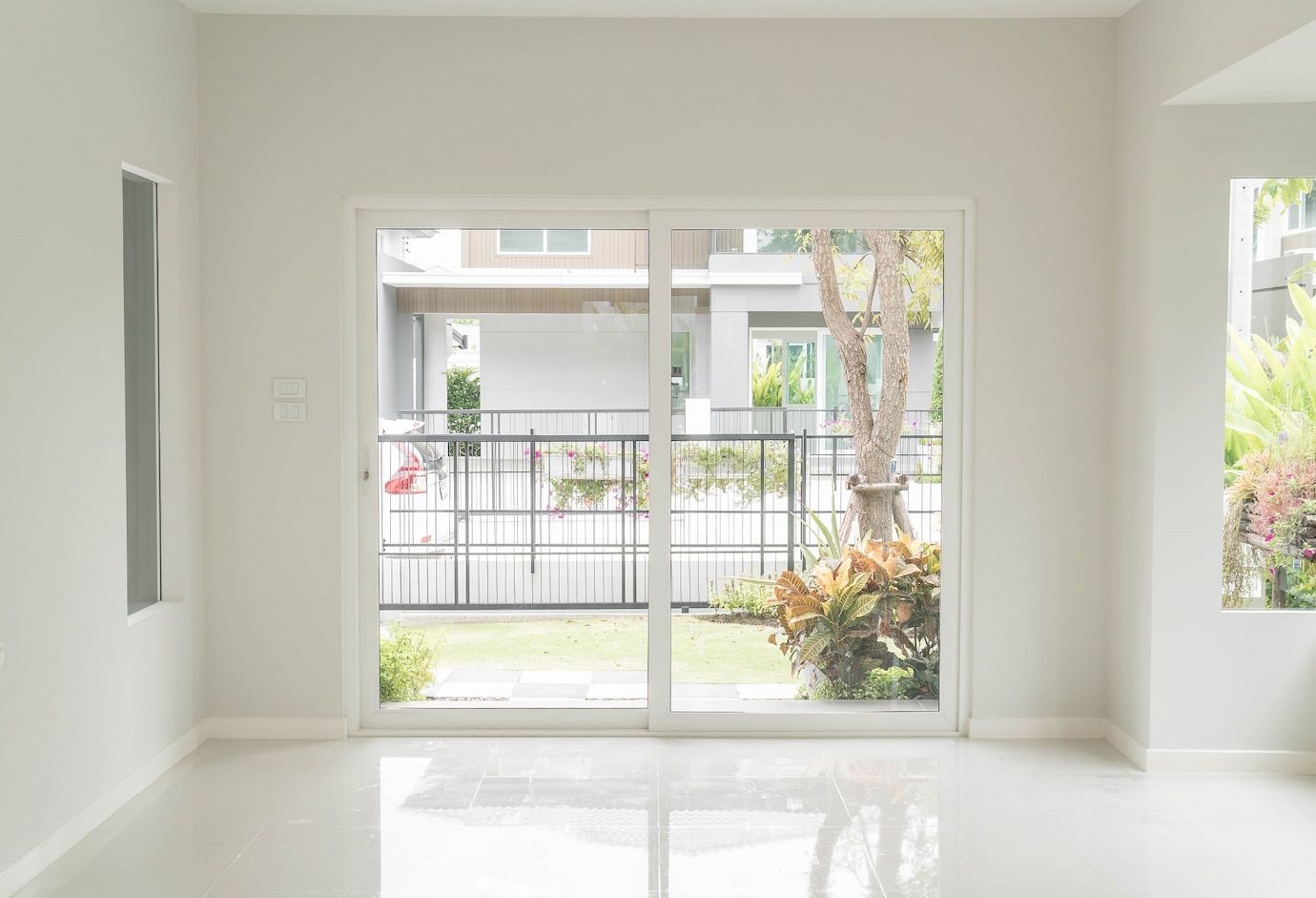 A bright, minimalist room featuring white walls, glossy tile floors, and a large glass sliding door looking onto a garden.